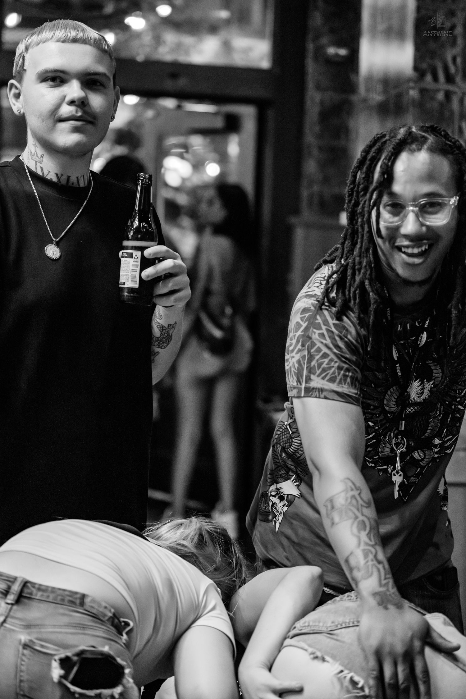 Two tattooed men laughing at a bar while a woman leans over in black and white.