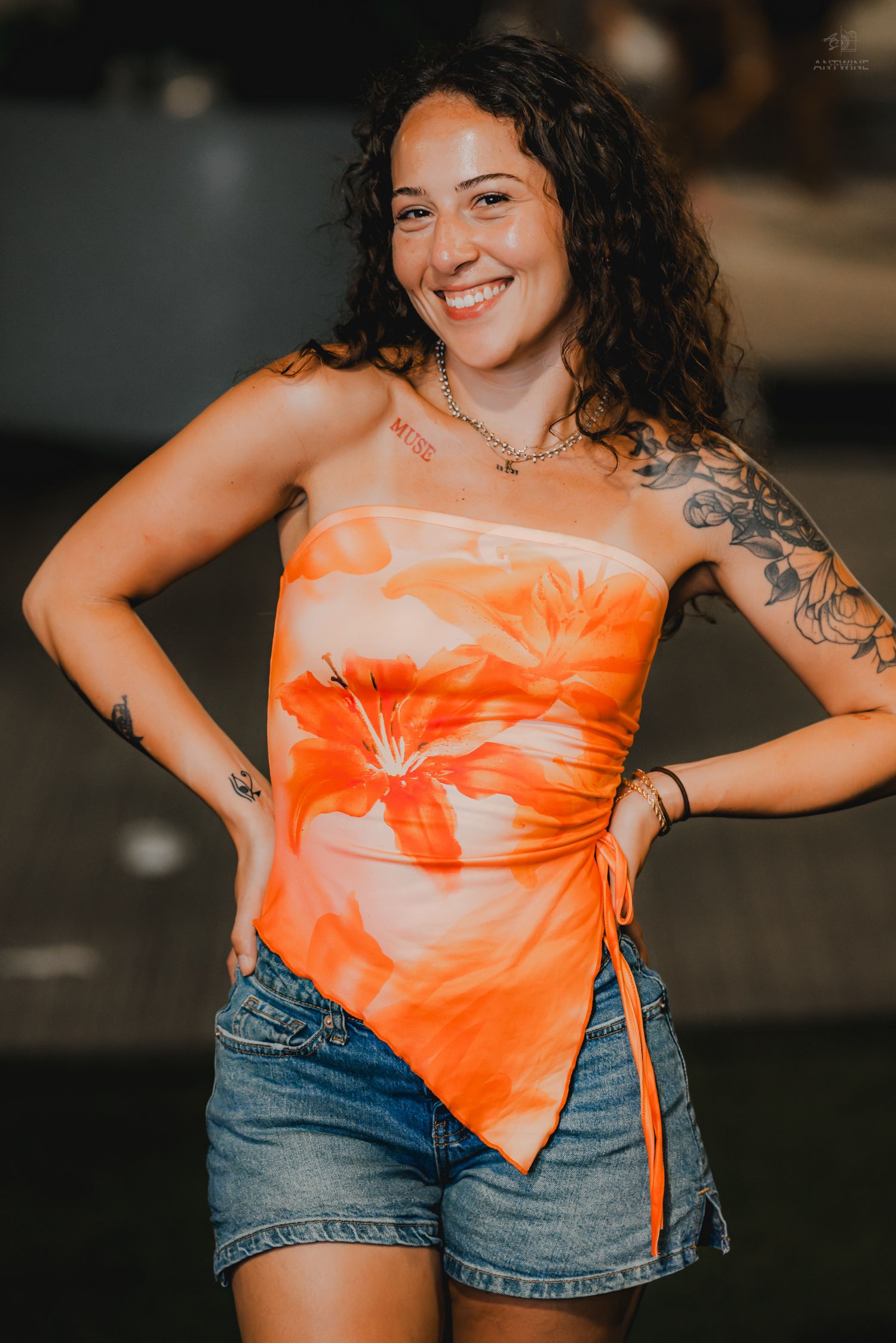 A tattooed woman in an orange floral strapless top and denim shorts smiles with hands on hips outdoors.