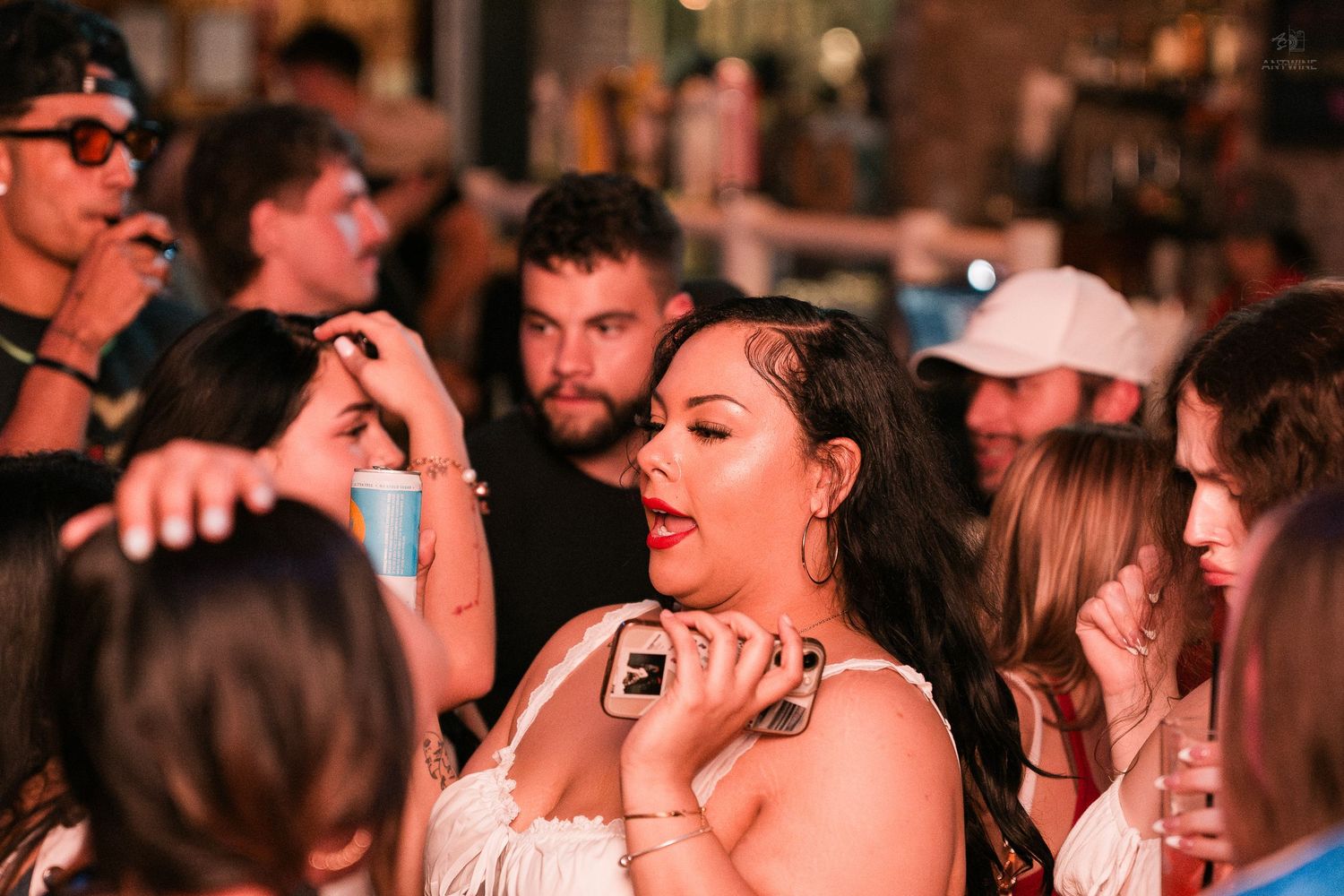 Woman in white dress holding a phone at a lively crowded bar event.