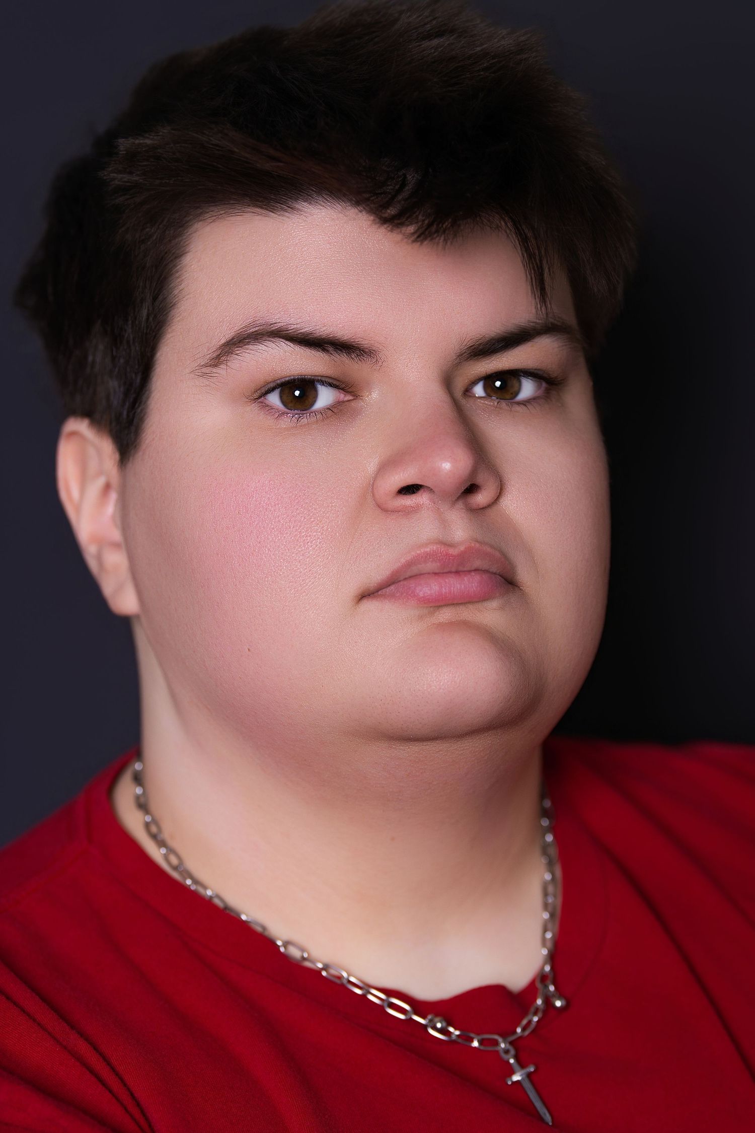 Close-up portrait of a young person in a red shirt wearing a silver chain necklace with a cross pendant.