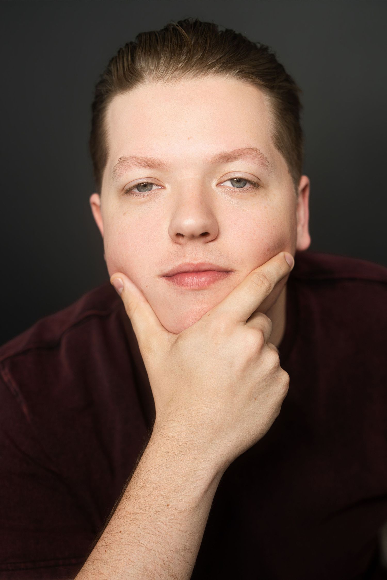 Color portrait of young man in maroon shirt resting chin on hand against dark gray background.