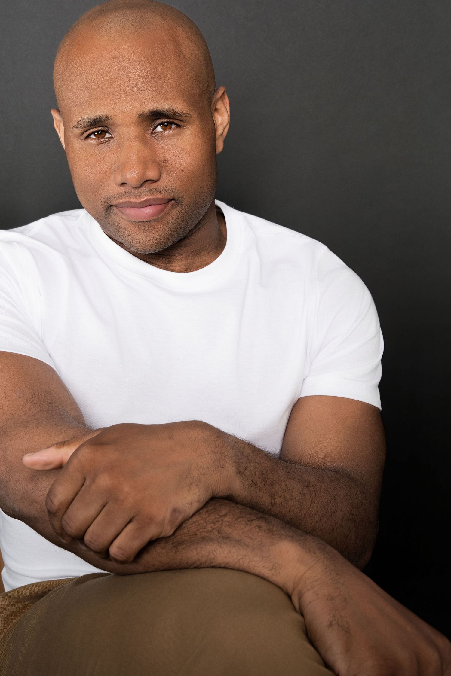 Confident bald Black man in white t-shirt sitting relaxed with arms crossed, looking at camera against dark background.