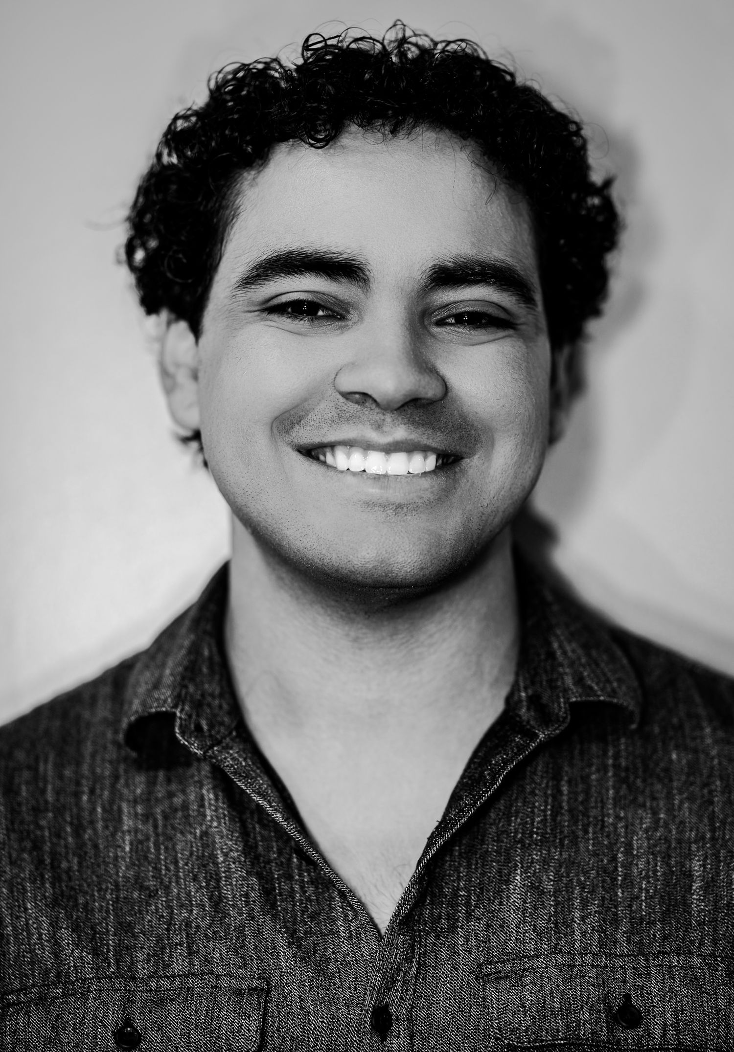Smiling young man with curly black hair wearing a dark shirt in a black and white headshot photo.