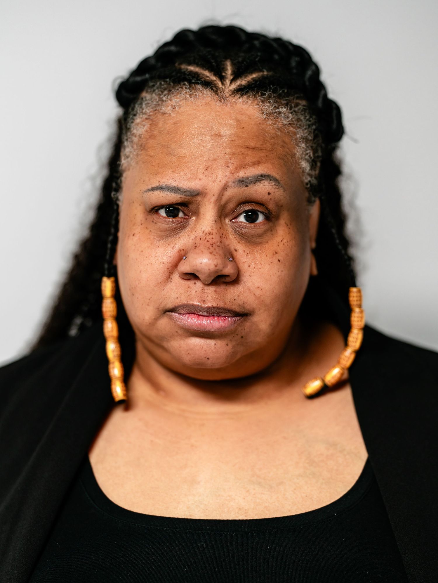 Woman with braided hair and gold earrings wearing black, with a serious expression, light background.