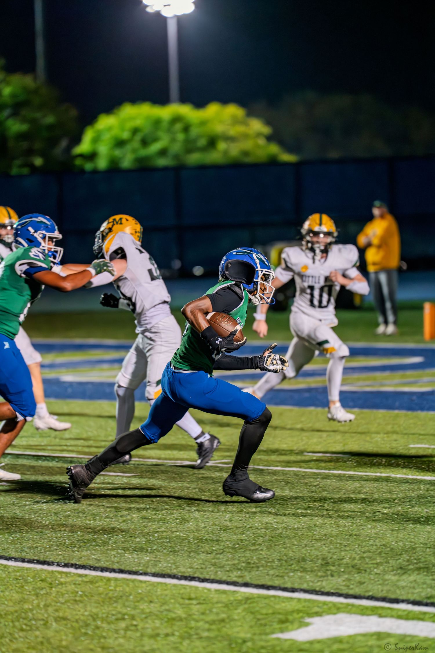 High school football player in green jersey breaks through tackle attempt during a night game.