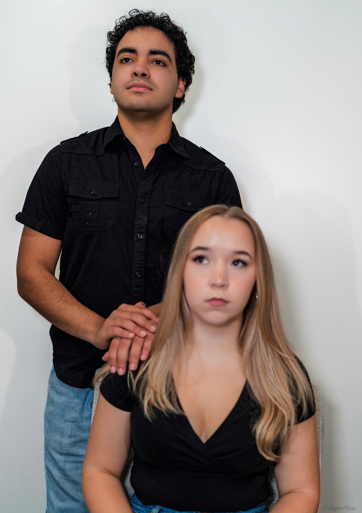 Young couple posing seriously, man standing behind seated blonde woman in color portrait.
