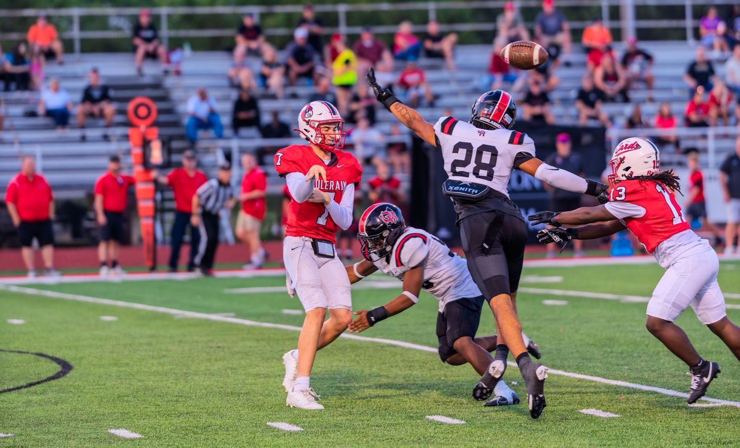 High school quarterback releases pass while defender leaps to block during nighttime football game.