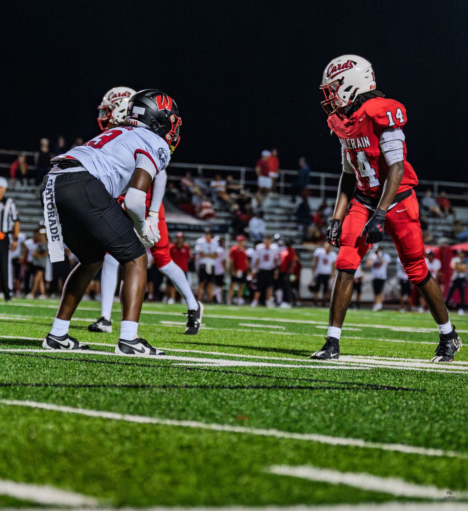 Two football players in red uniforms face off on turf field during nighttime high school game.