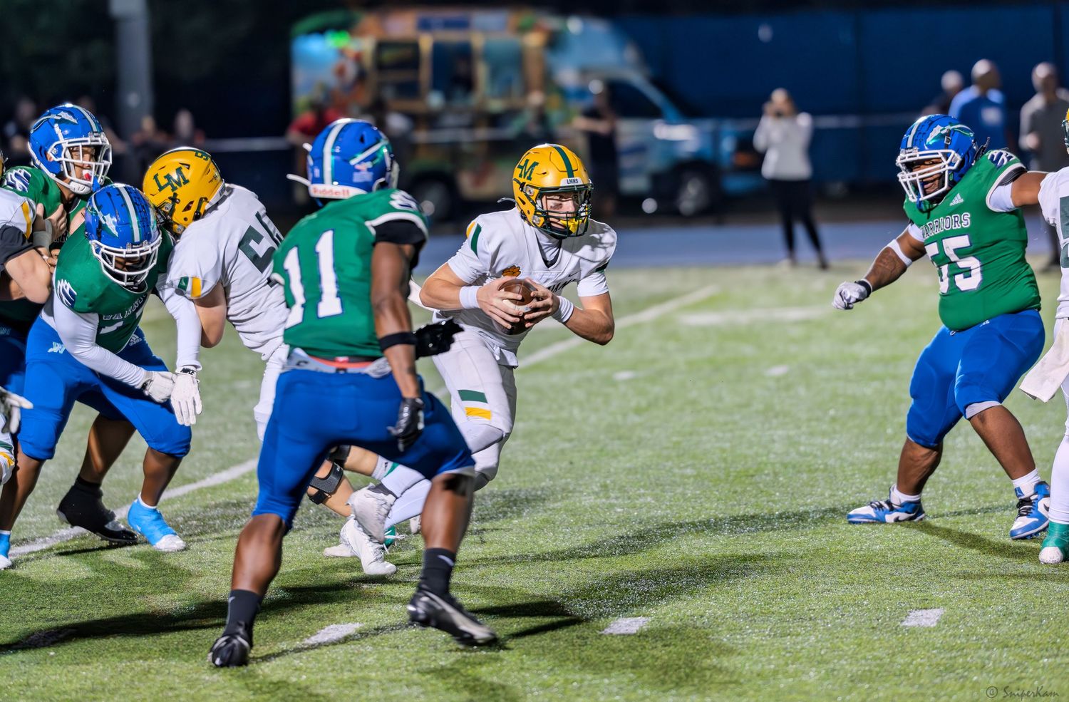 High school football player in gold and white uniform runs through defenders in blue and green jerseys.