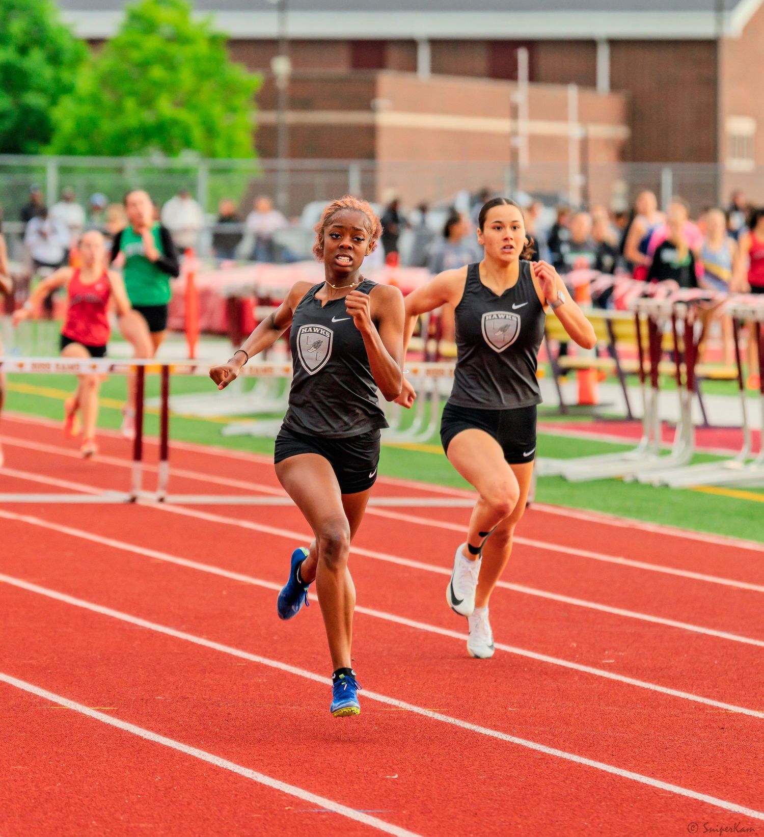 Two female athletes in matching black uniforms sprint toward the finish line on a red track.