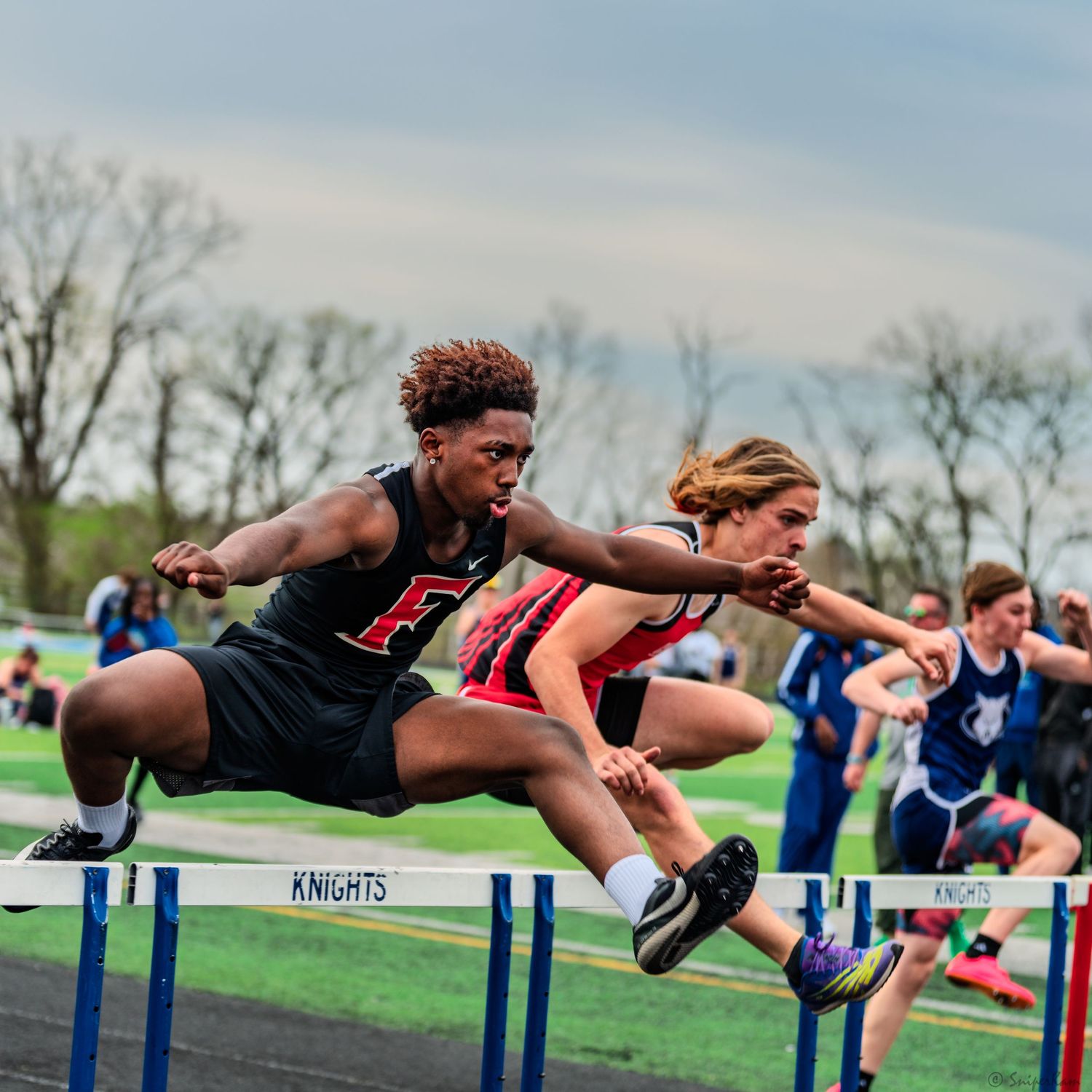 Male athlete leaping over hurdles during a competitive high school track and field meet.