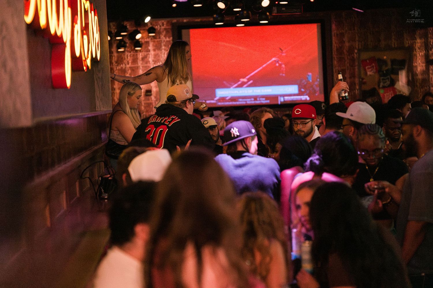 Packed sports bar crowd watches game on large projection screen under red neon lights.