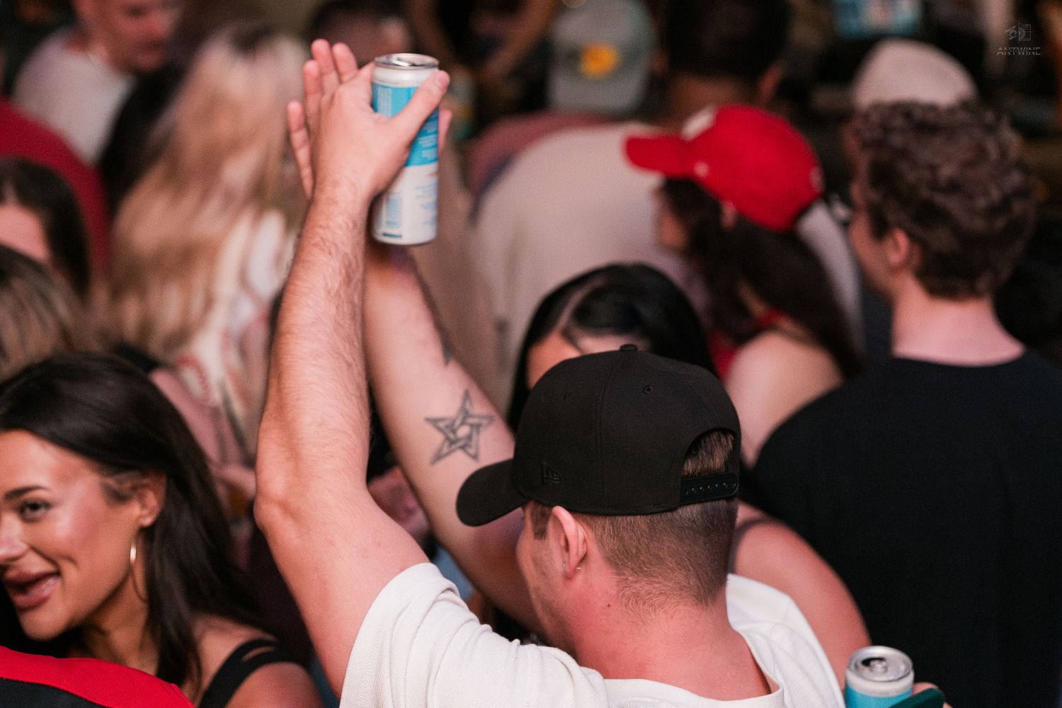 Man in black cap raising a beer can above the crowd at a packed bar.