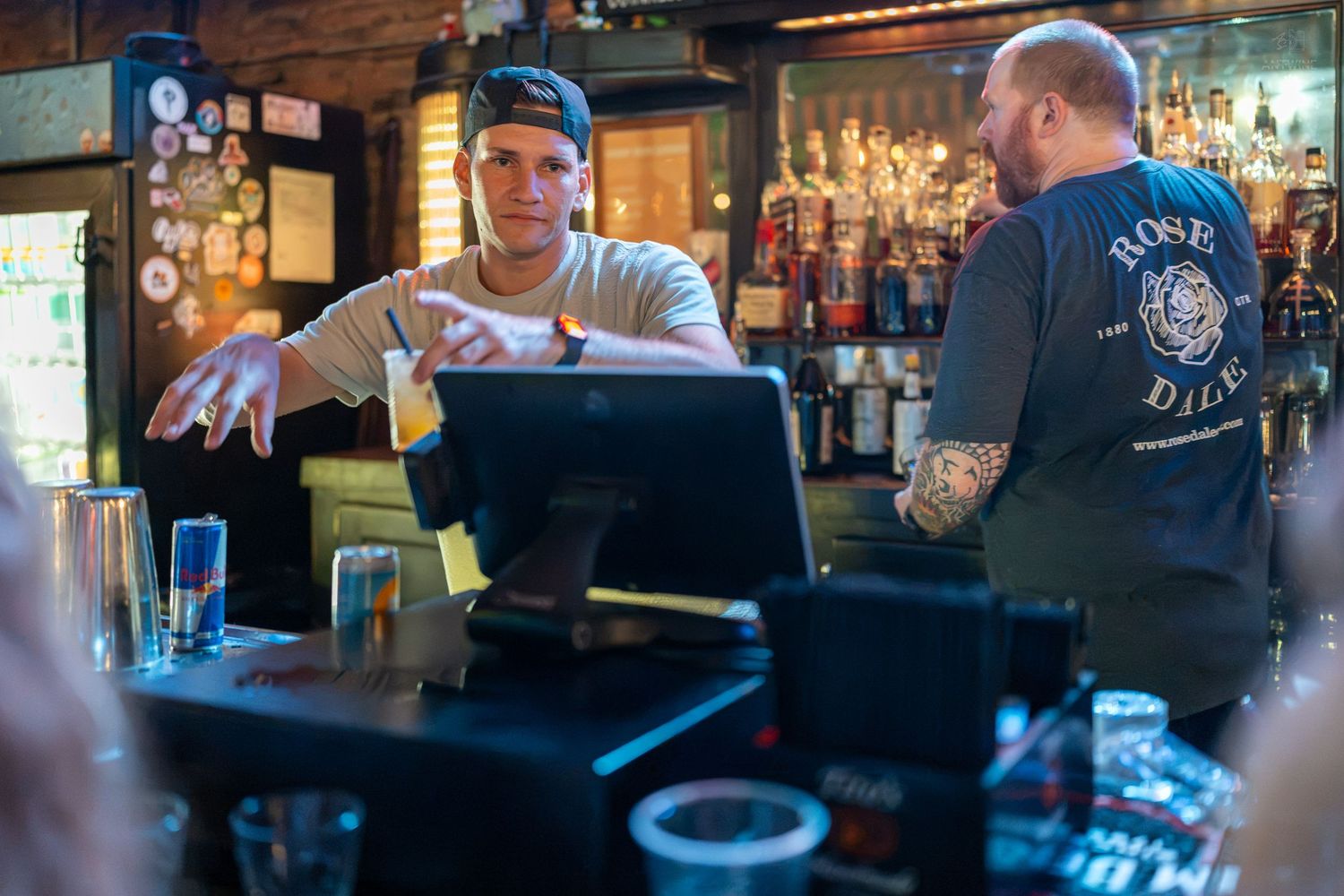 Bartender and customer converse at a rustic bar with liquor bottles and a POS system on the counter.