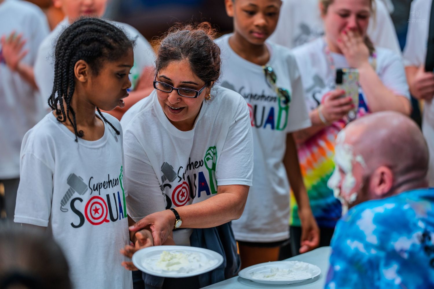 Children and adults in Superhero Squad shirts participate in a pie-eating contest at a fun outdoor event.