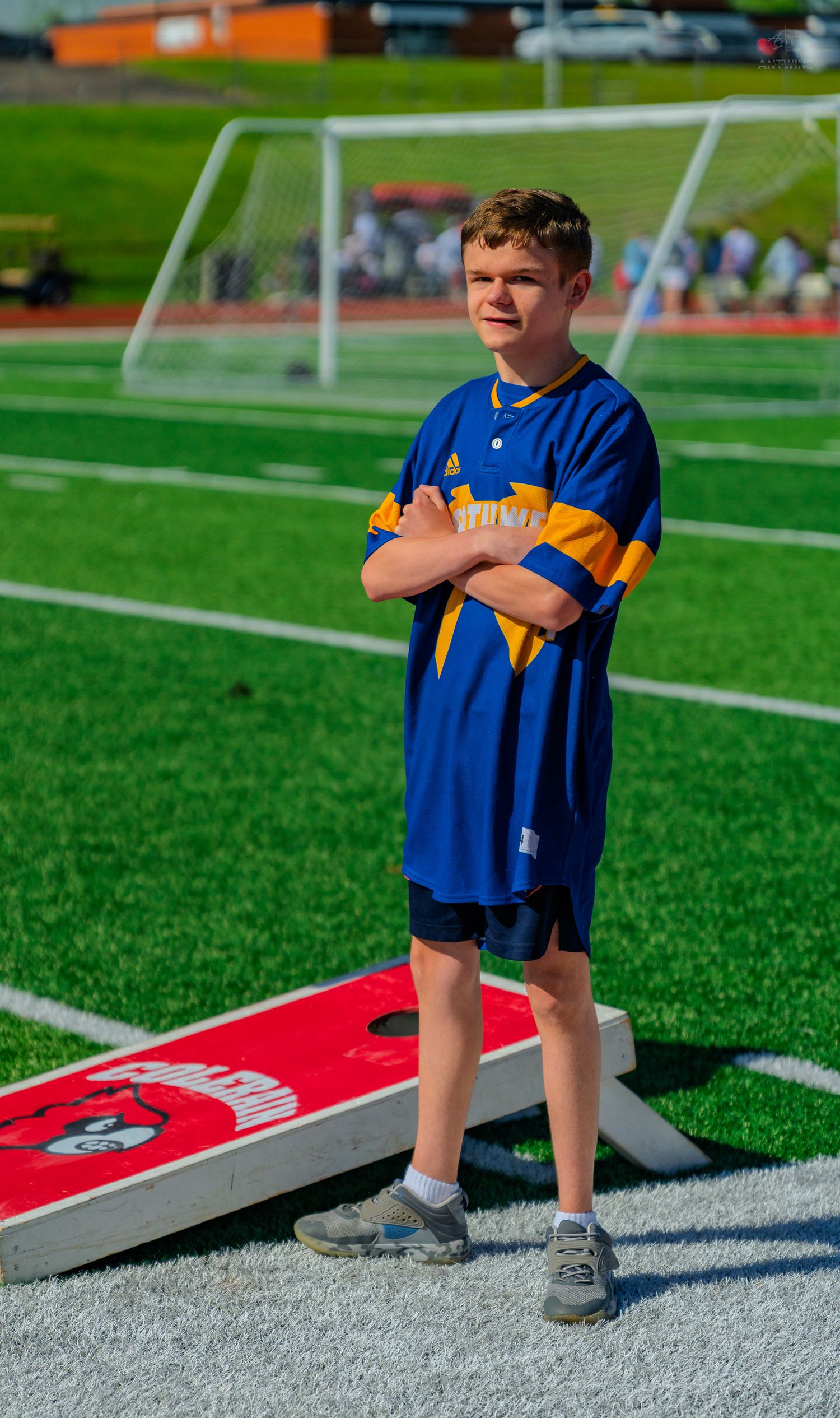Boy in blue Adidas jersey stands arms crossed on green turf field next to red cornhole board with soccer goal behind.