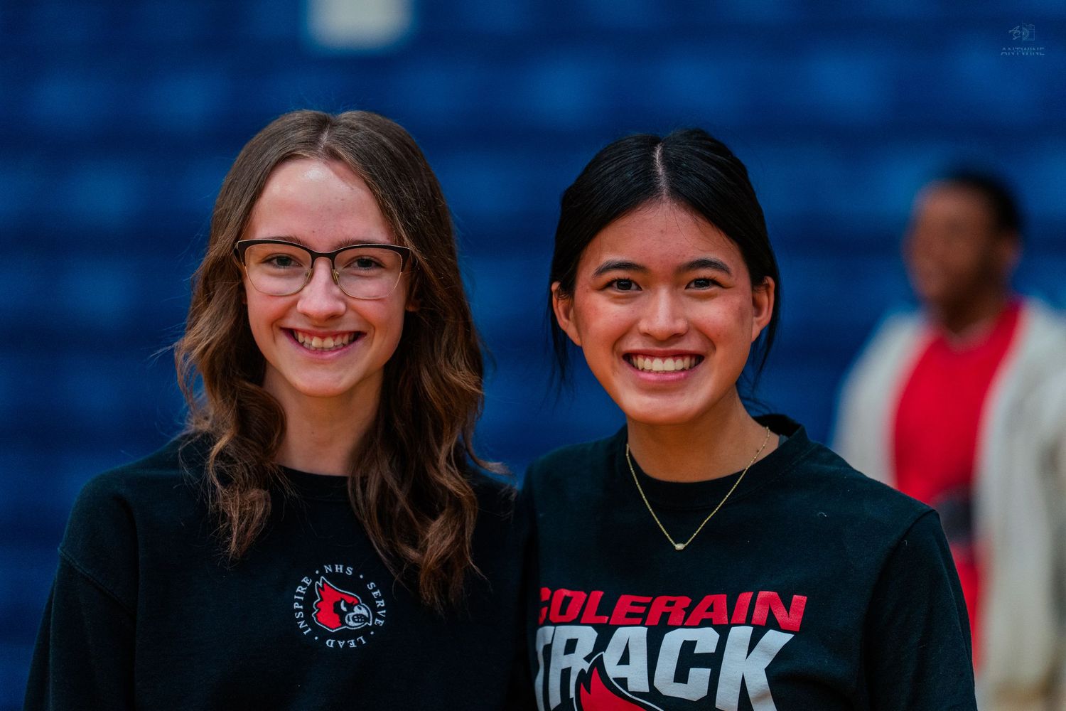 Two smiling high school girls in Colerain Track shirts pose together in a gymnasium.