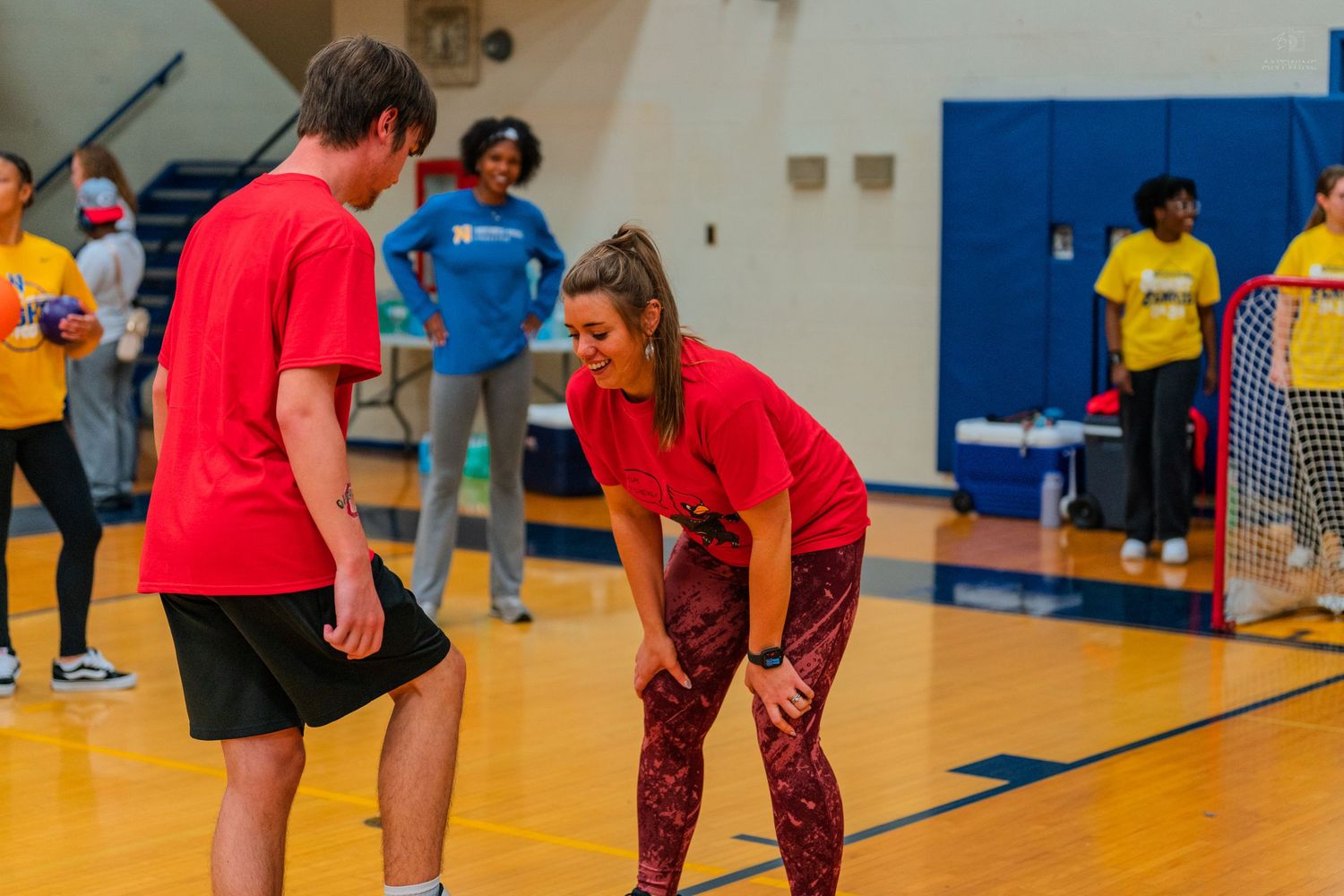 Two people in red shirts laugh together on a gym court during an indoor sports activity event.