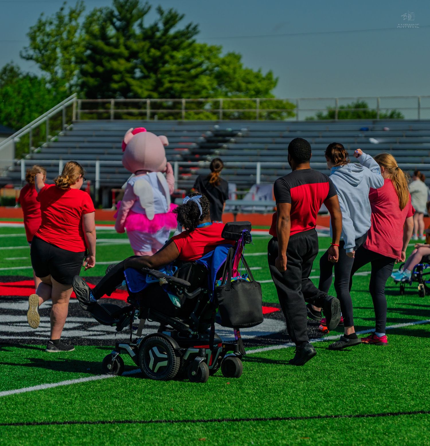 Participants including a person in a power wheelchair walk across green turf at an inclusive sports event.