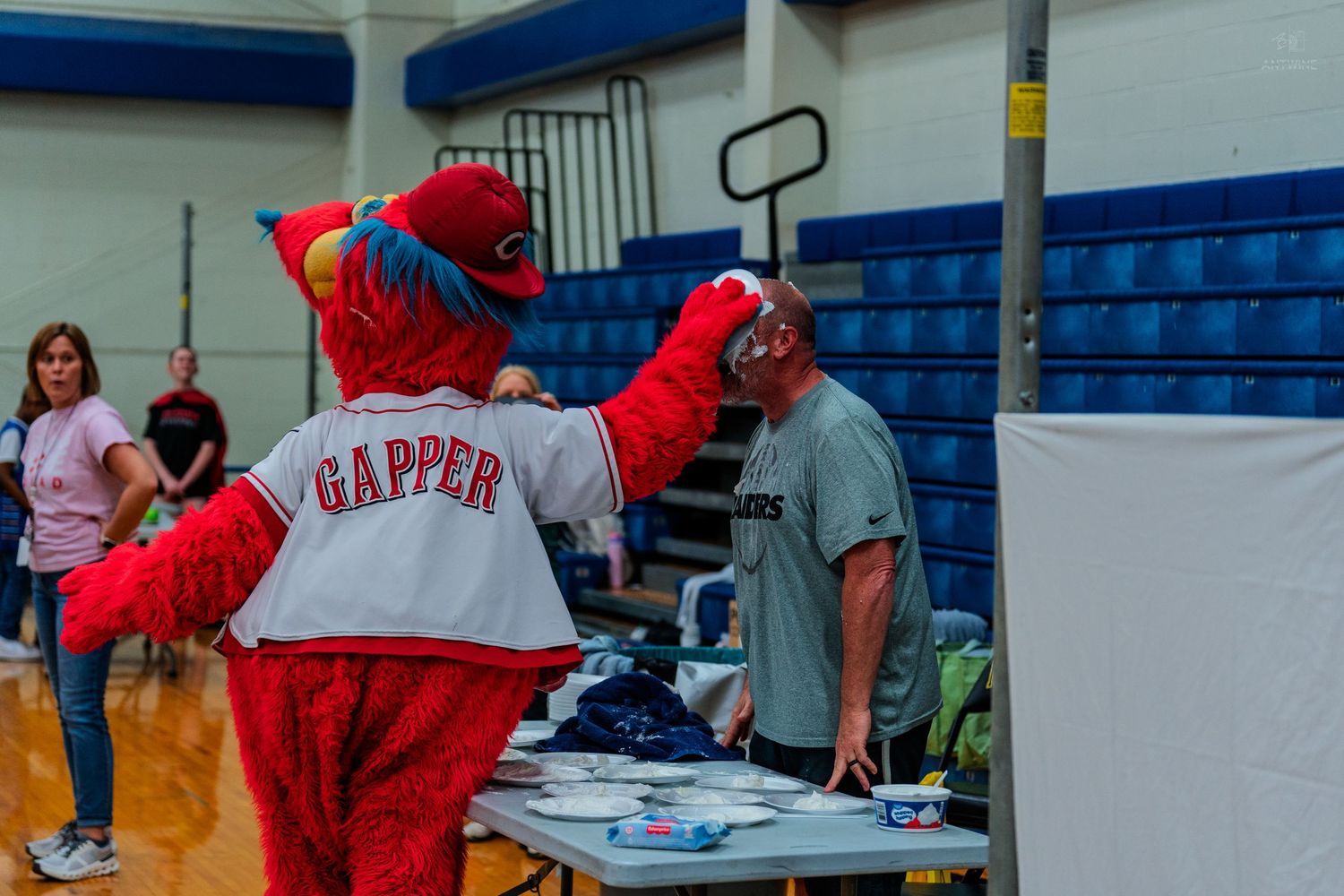 Gapper, the Cincinnati Reds mascot, interacts with a volunteer at an indoor event table in a gymnasium.