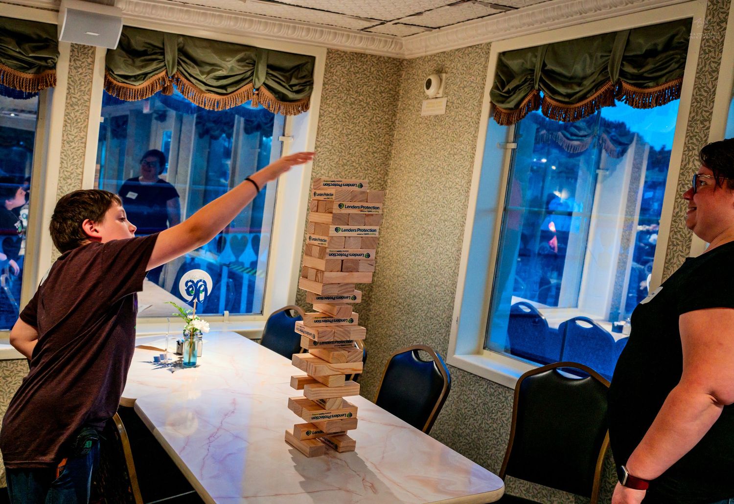 Two people playing giant Jenga at an indoor table, one player reaching to remove a wooden block from the tall tower.