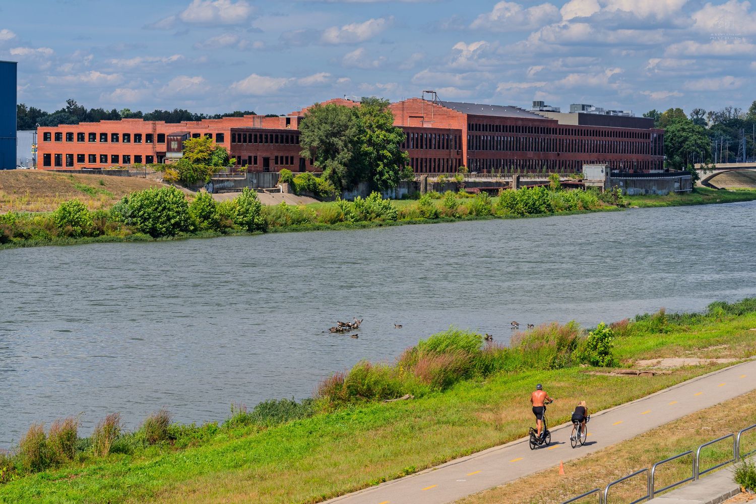 Cyclists riding along a riverside path with red brick buildings across the water on a sunny day.