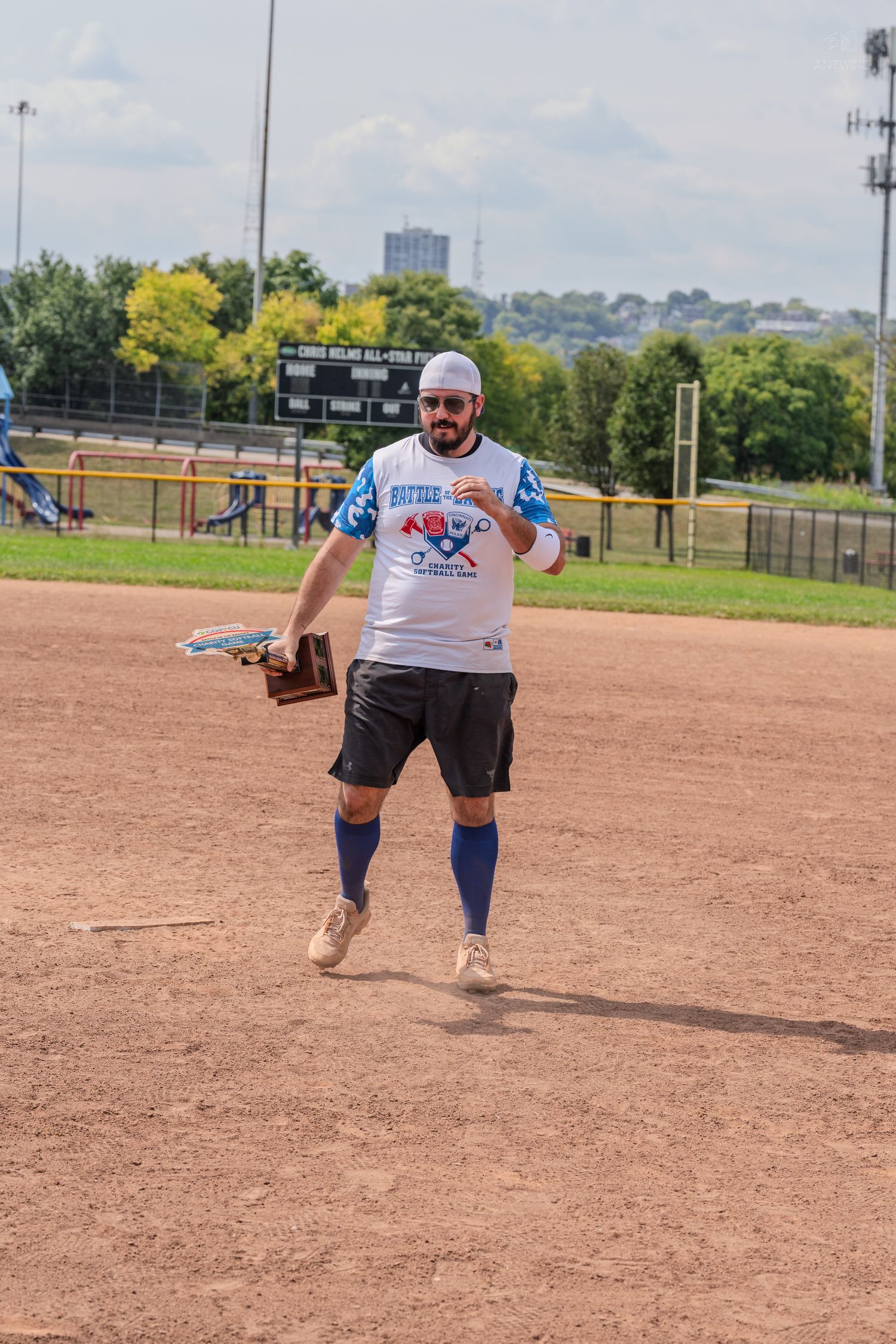 A bearded man in a white softball jersey and blue knee socks runs across a dirt baseball field during a charity game.