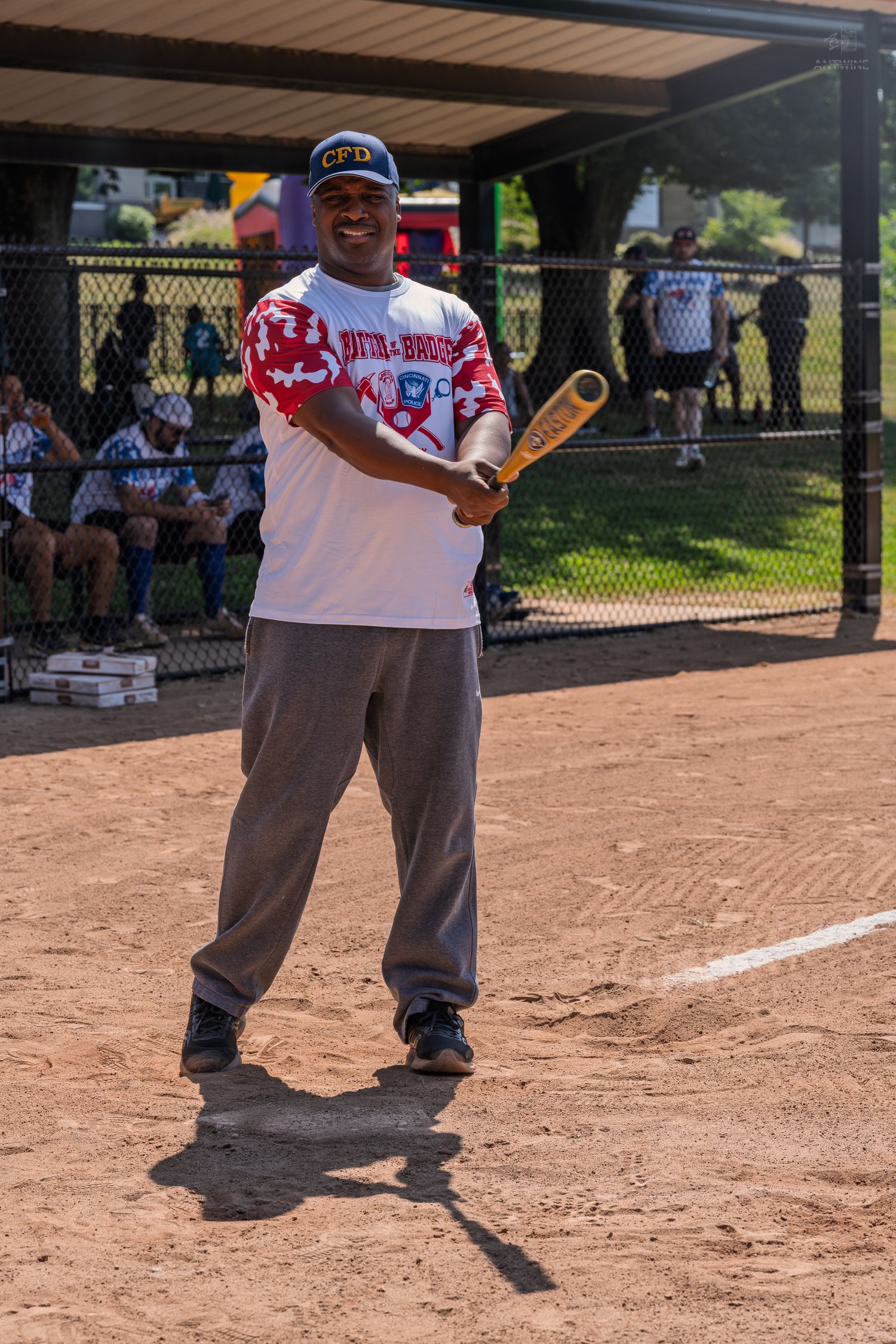 A smiling baseball player poses with a bat at home plate during a sunny outdoor softball game.