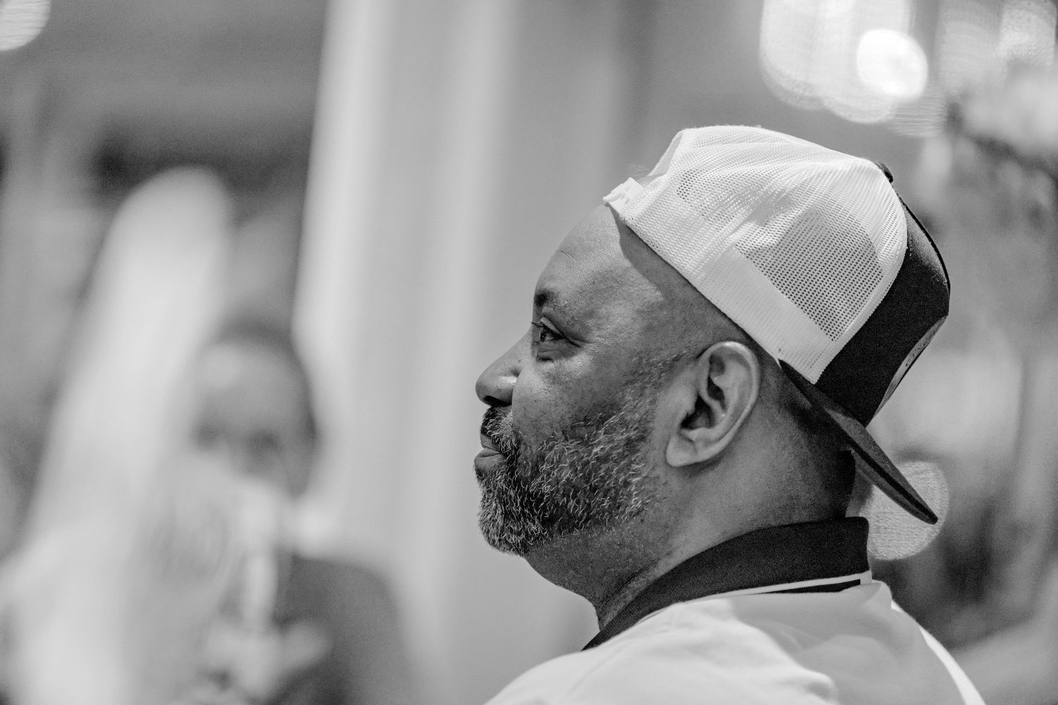 Black and white profile portrait of a bearded man wearing a snapback cap indoors.