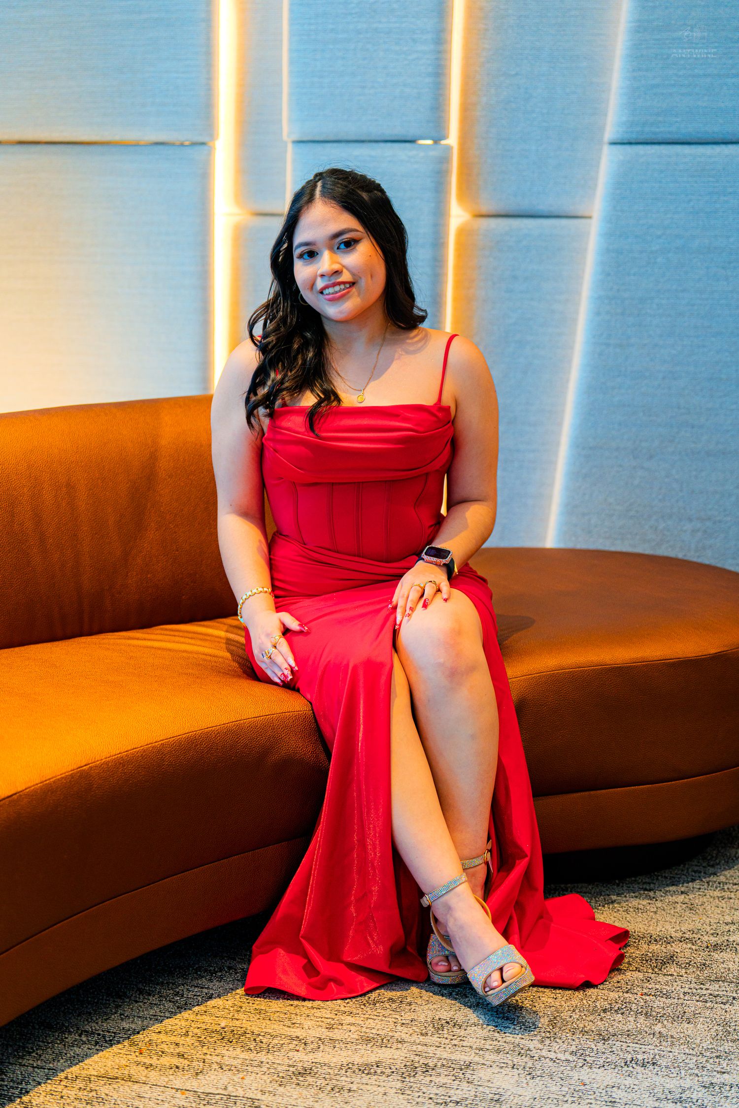 Young woman in elegant red satin gown sitting gracefully on an orange curved sofa in a modern interior.
