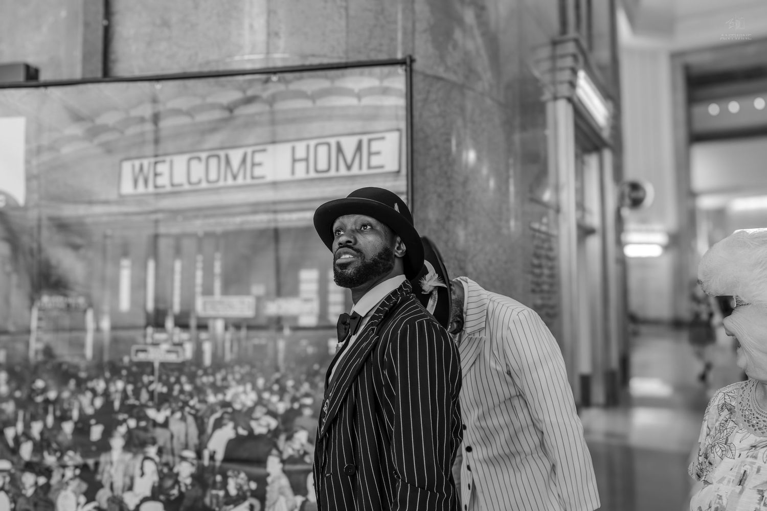 Black and white photo of a man in a pinstripe suit standing before a Welcome Home sign and vintage crowd mural.
