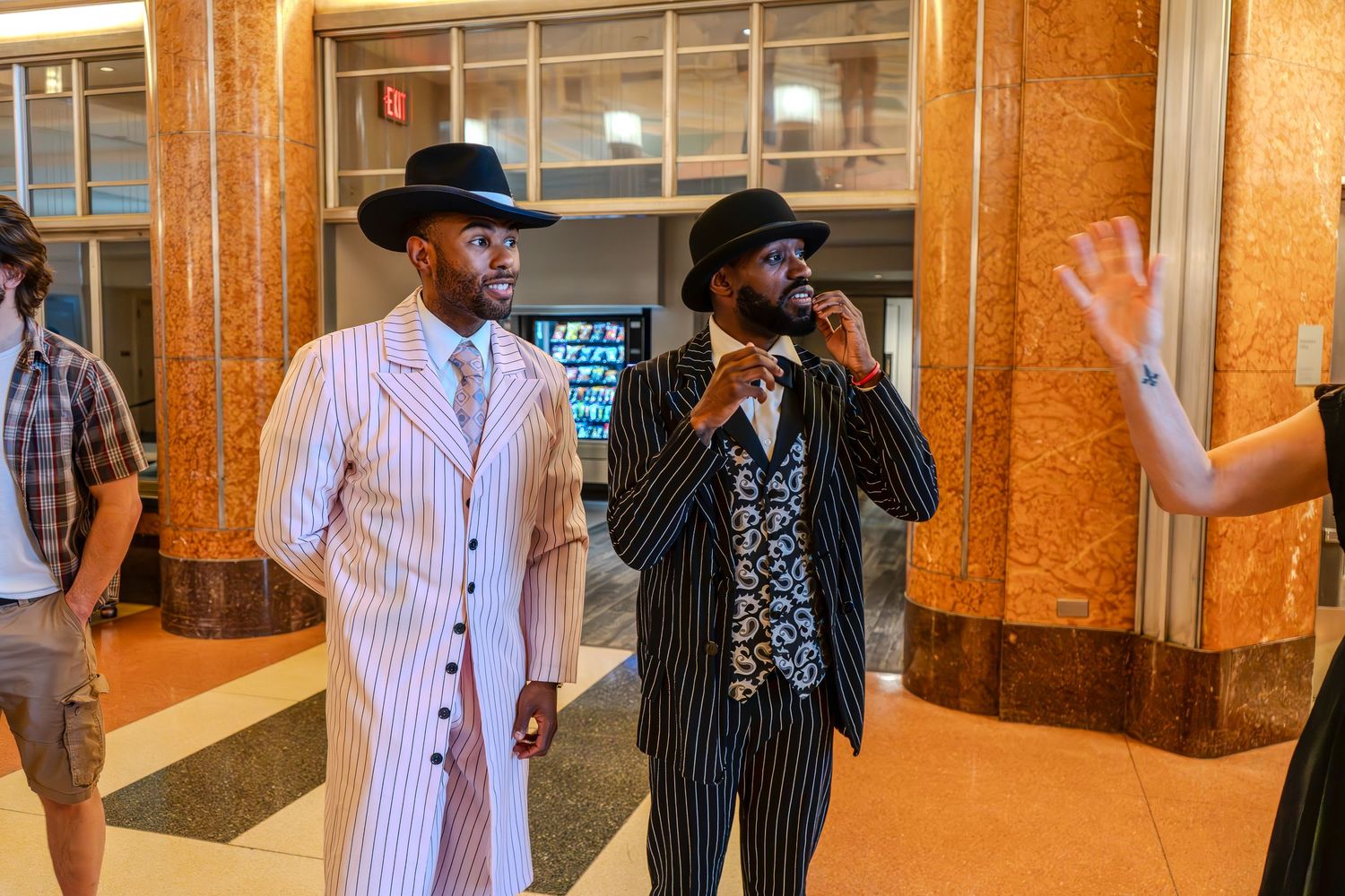 Two men in stylish pinstripe zoot suits and fedora hats stand in a marble lobby.