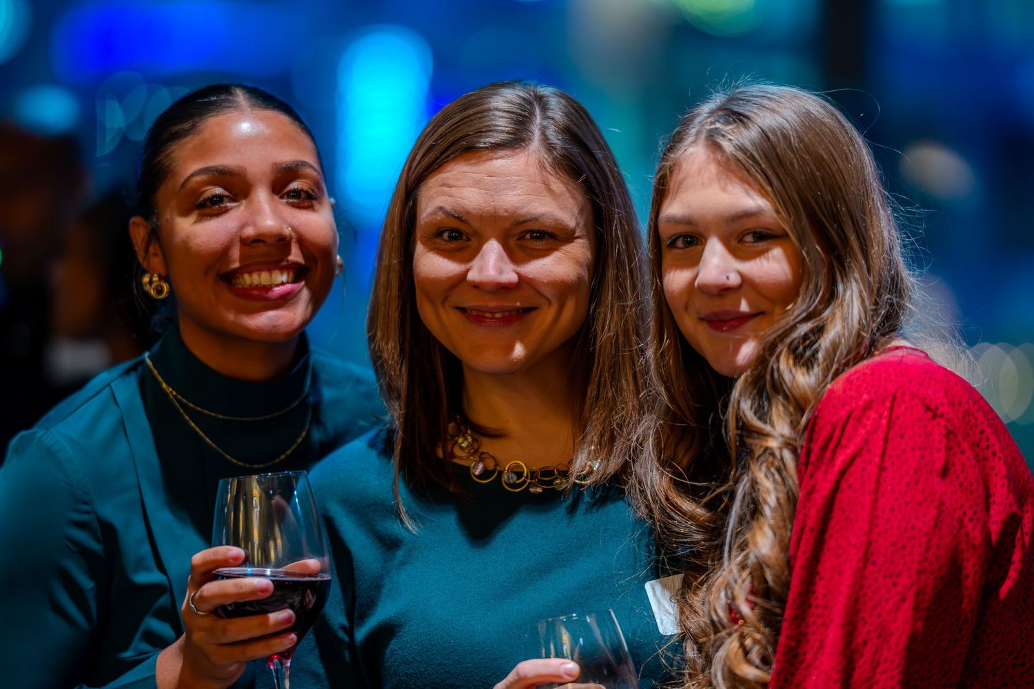 Three smiling women posing together at a social event, holding wine glasses, dressed in teal and red outfits.