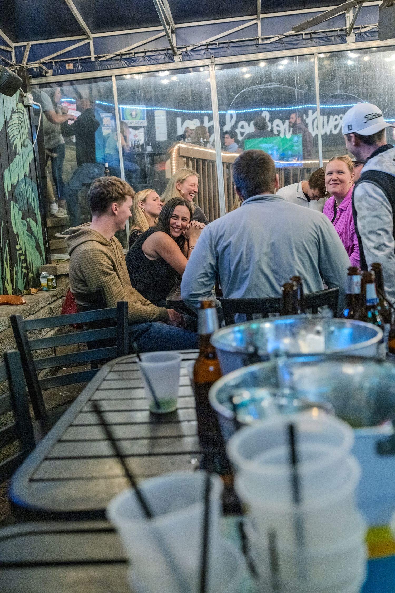 Friends socializing at an outdoor bar table with beer bottles and ice buckets at a night event.