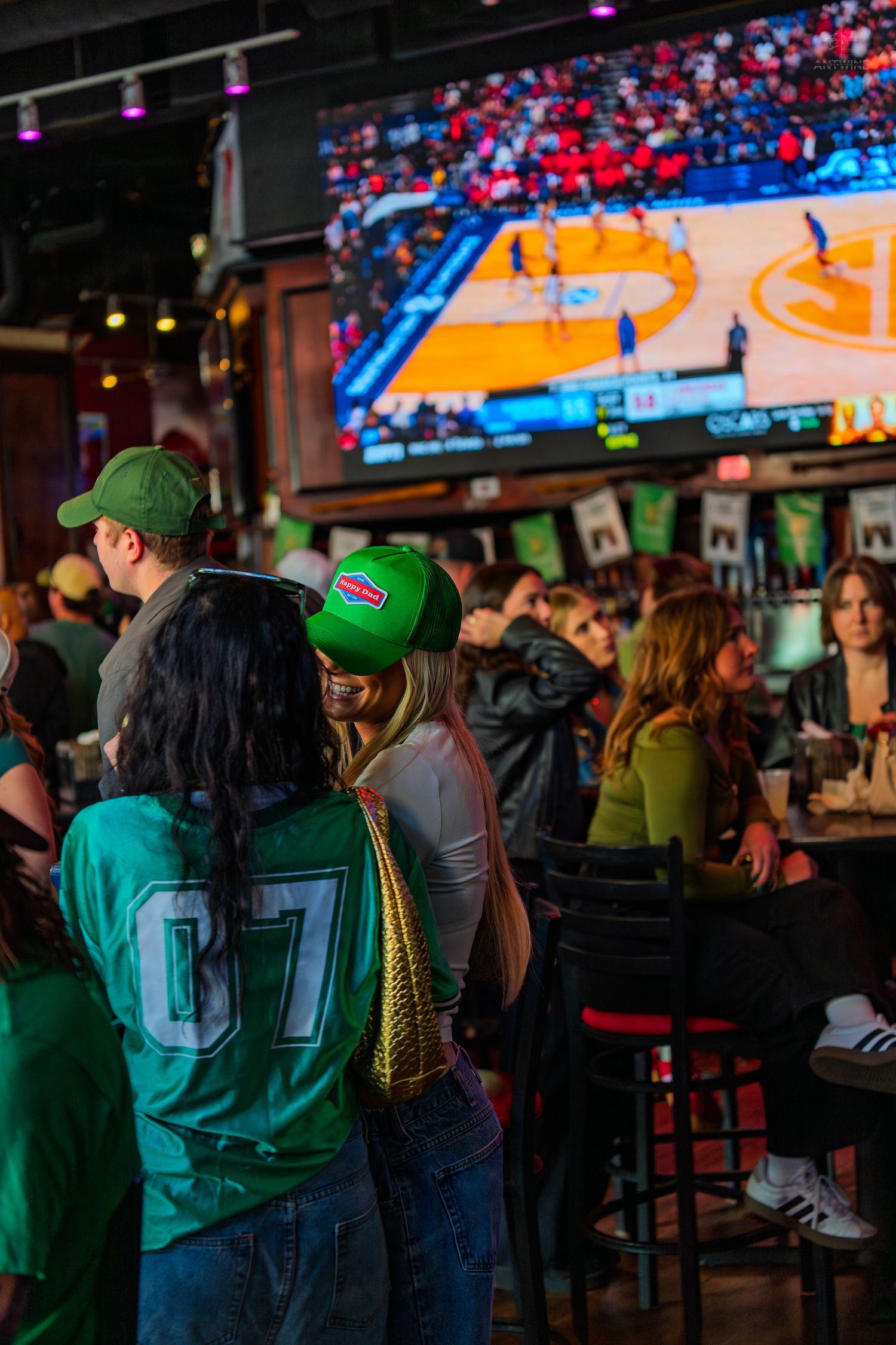 Fans in green jerseys and hats watch basketball on a big screen at a lively sports bar.