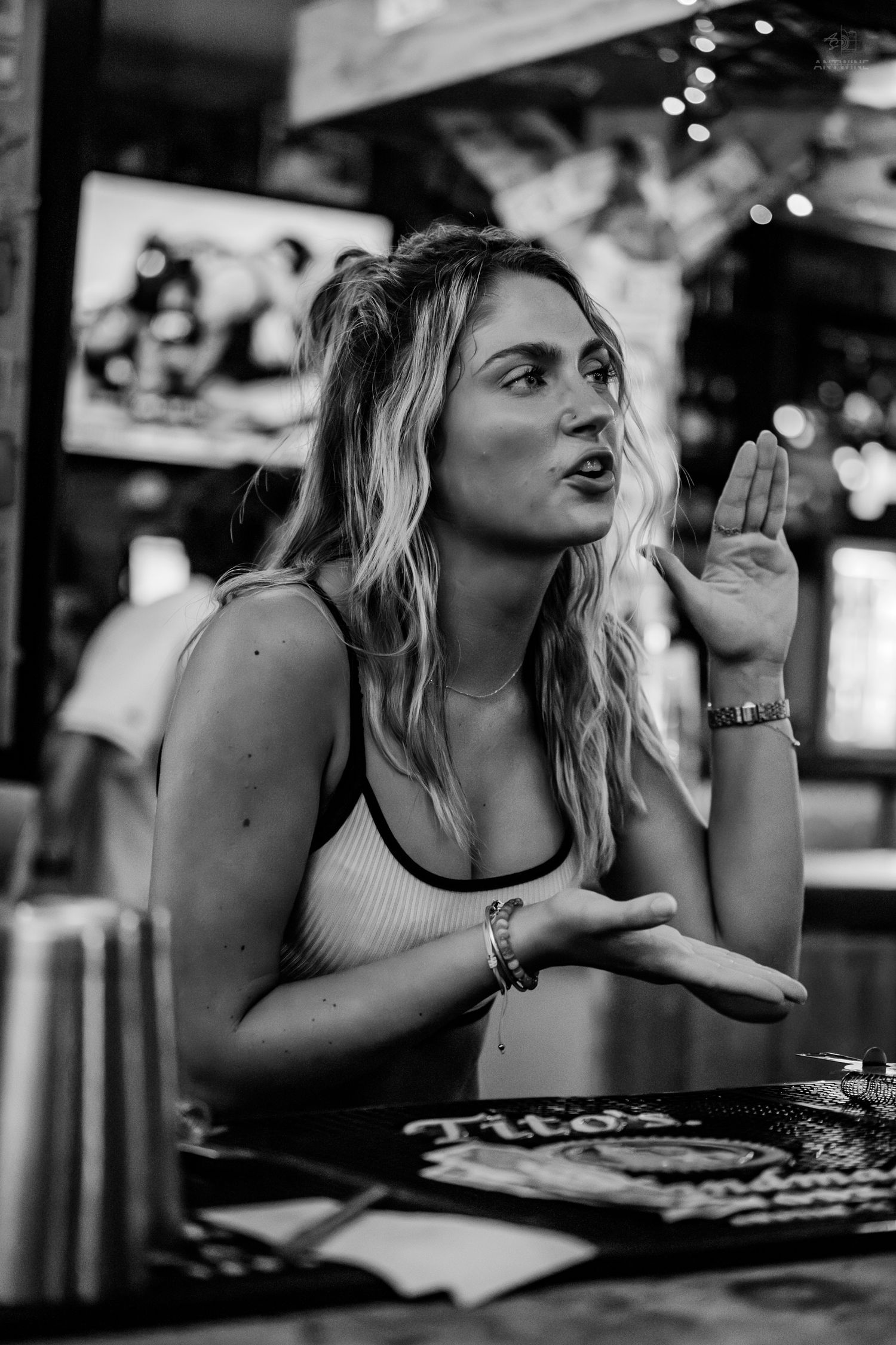 Black and white photo of a blonde woman gesturing while talking at a bar table.