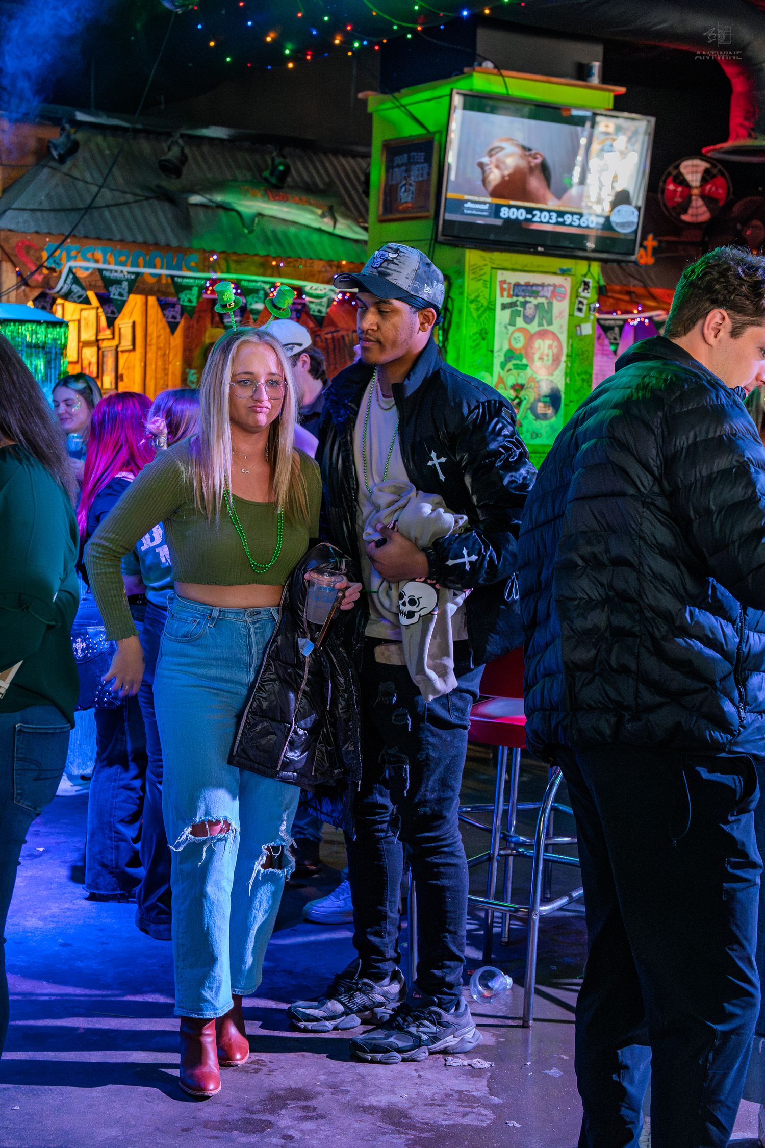 Couple posing at a vibrant nightclub, woman in ripped jeans and green top, man in black leather jacket.