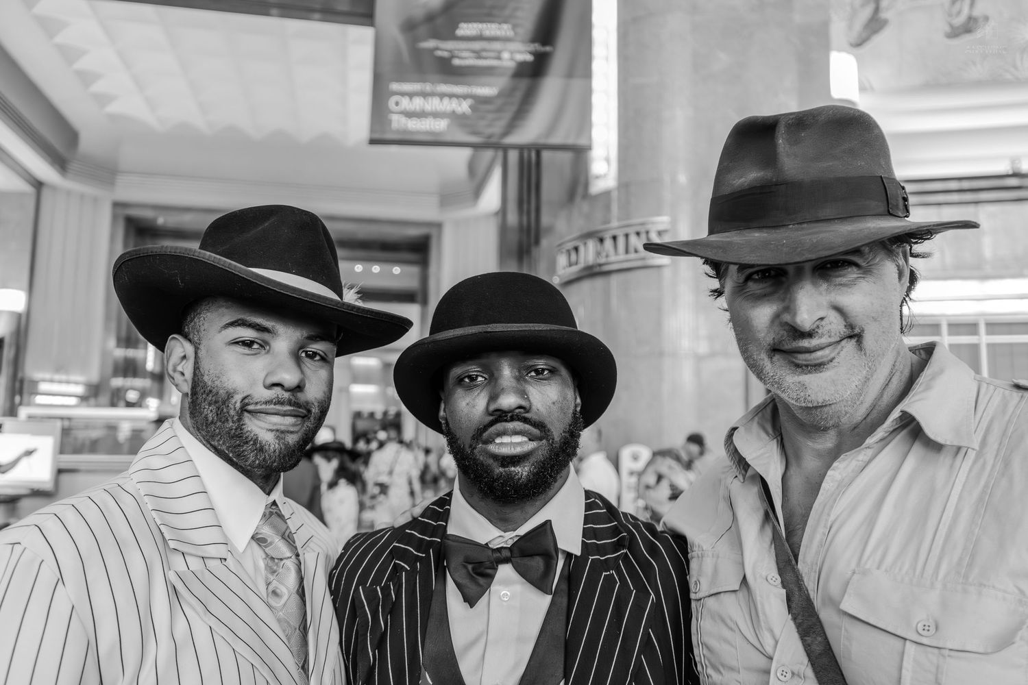 Three stylish men wearing fedora hats pose together in a black and white photo at a formal venue.