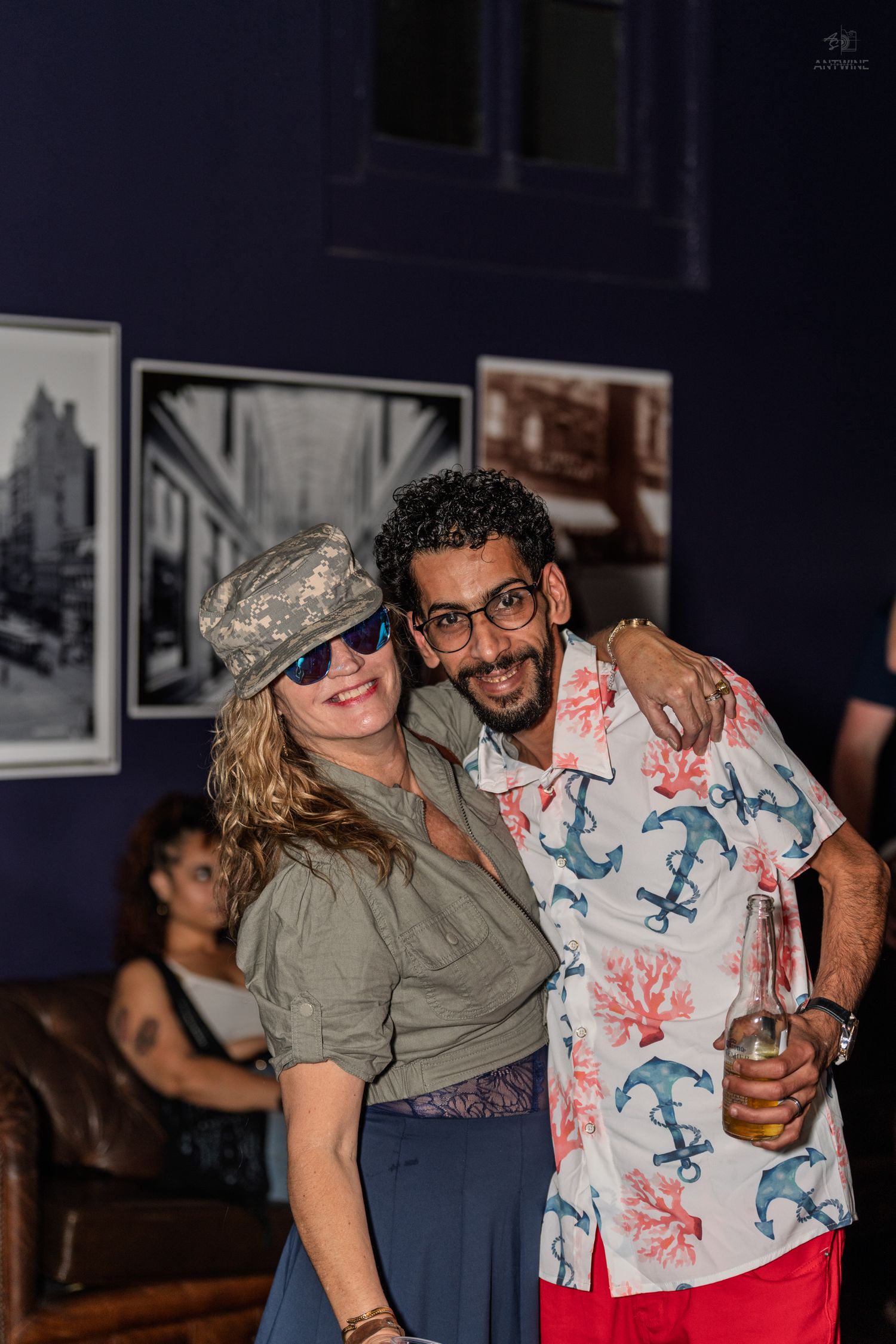Two friends pose together at an art gallery event, woman in camo hat and sunglasses with man in floral shirt.
