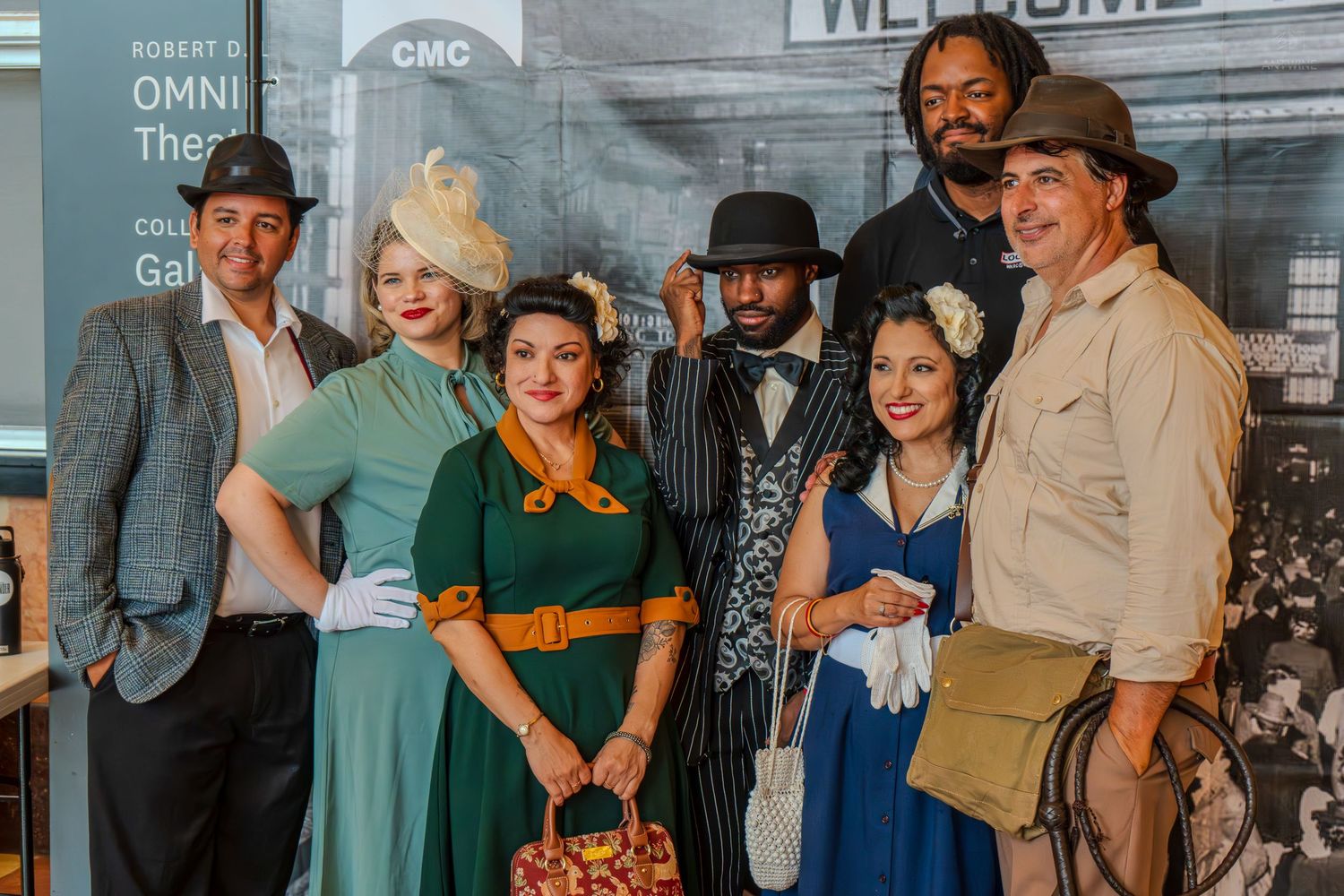 Group of seven people in 1920s vintage costumes posing together at the Omni Theater CMC event.