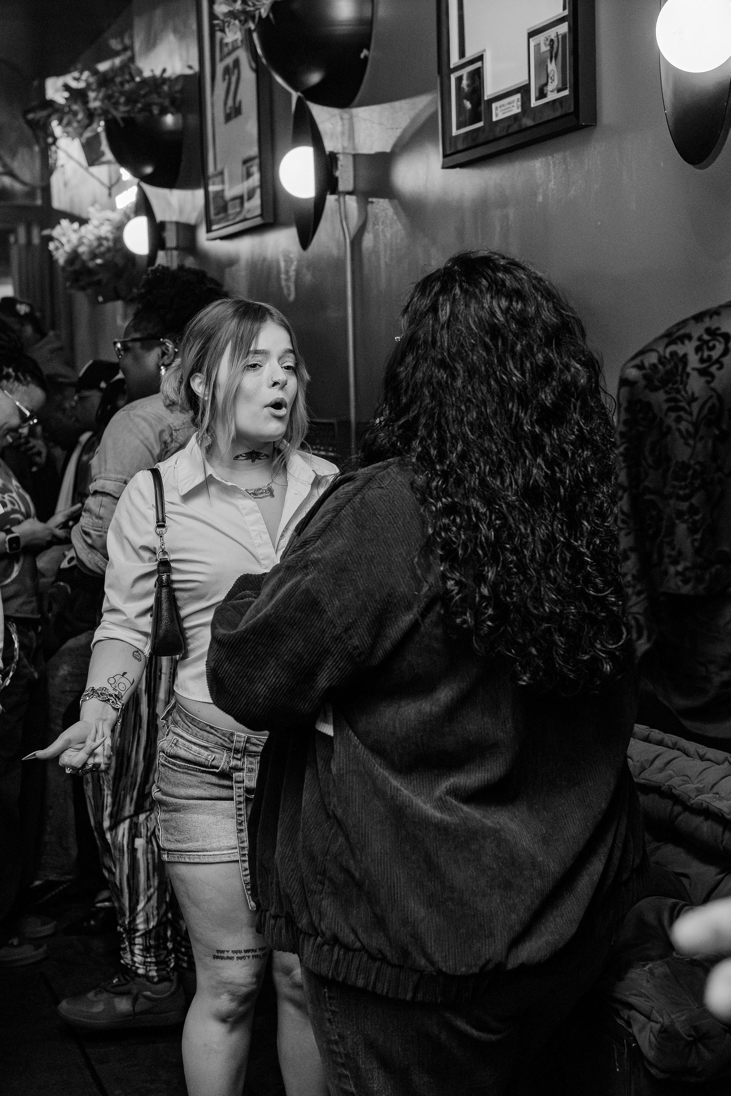Two women conversing in a crowded bar, captured in black and white photography.