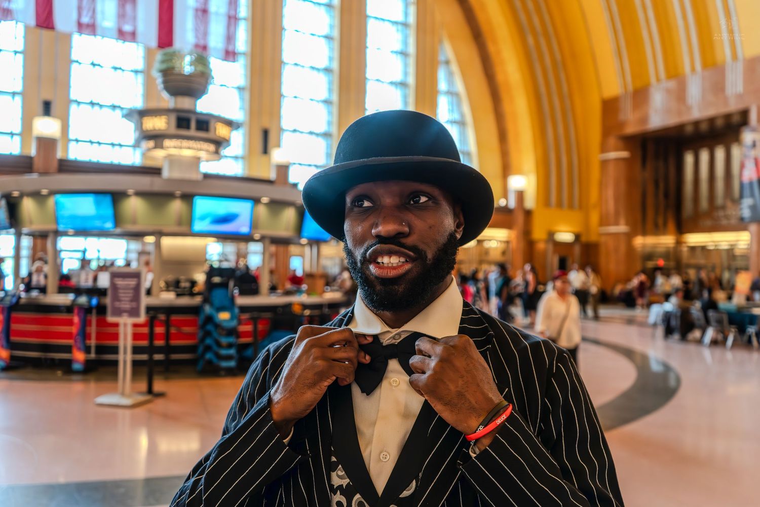 Stylish man in pinstripe suit and bowler hat adjusting bow tie inside a grand train station.