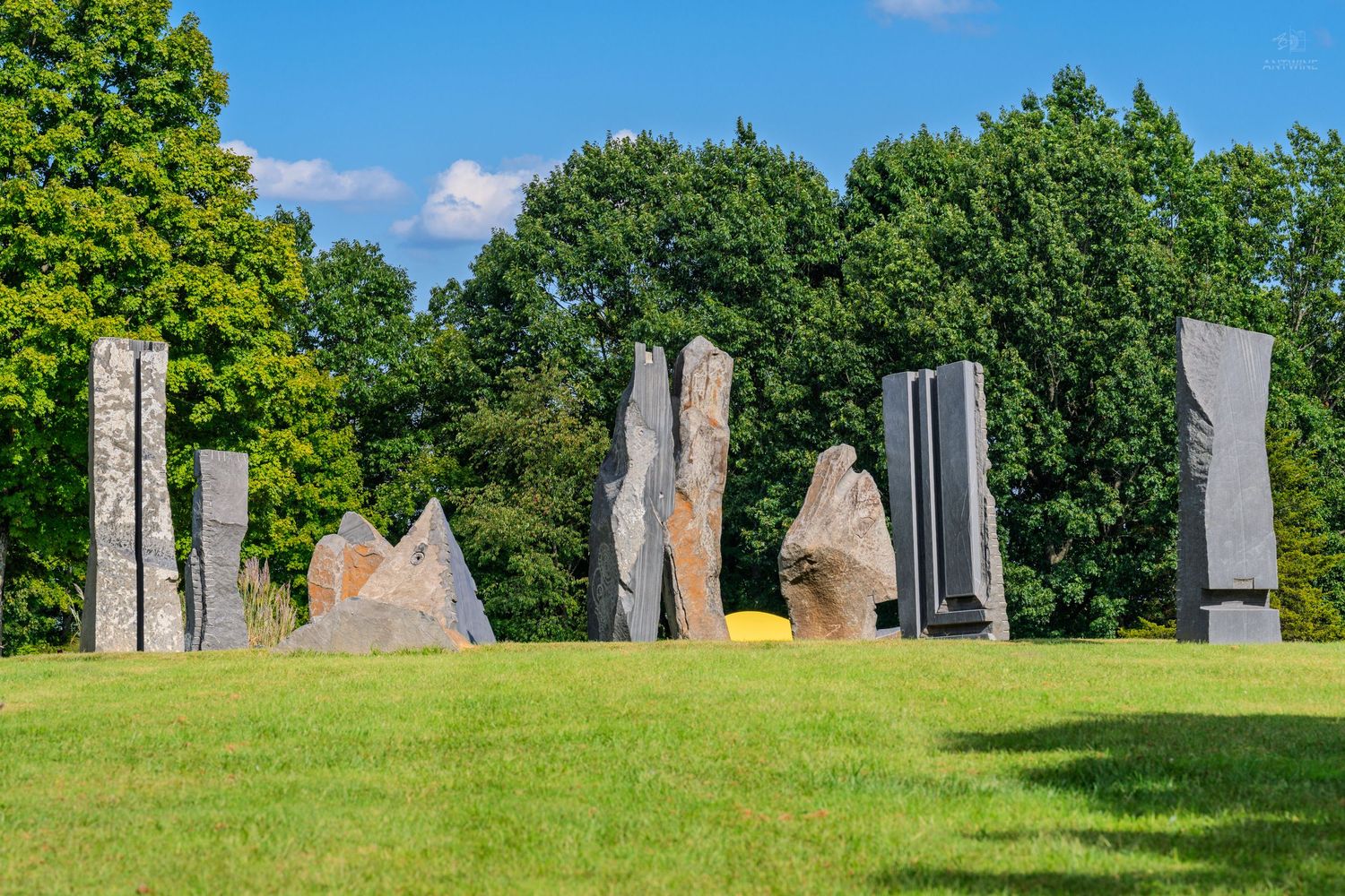 Large stone sculptures standing upright on green grass, surrounded by tall trees under a bright blue sky.
