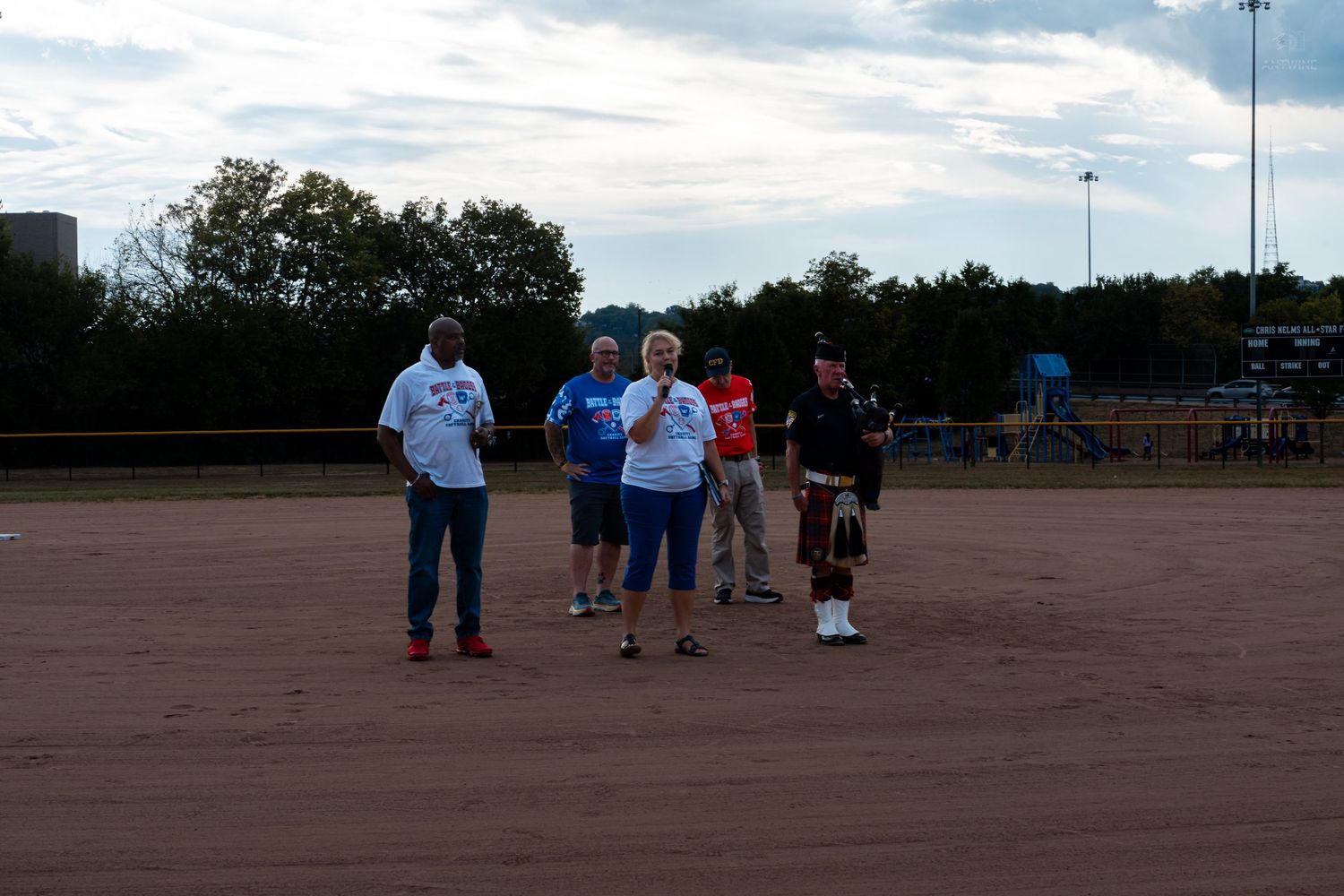 Small group of people including a bagpiper standing on a dirt softball field at dusk.