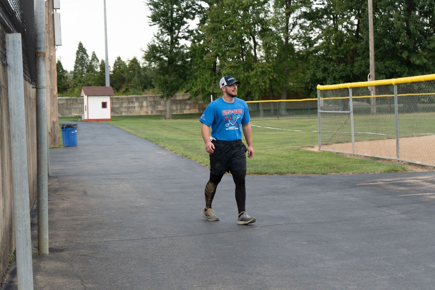 Man in blue t-shirt and black pants playing baseball near a field on a paved path.