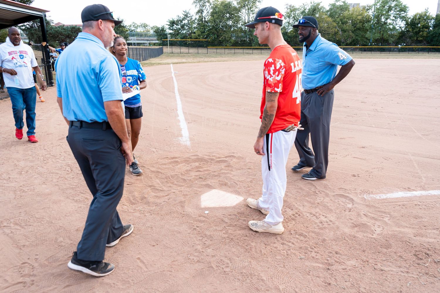 Baseball players and umpires meet at home plate on a dirt field during a softball game at dusk.
