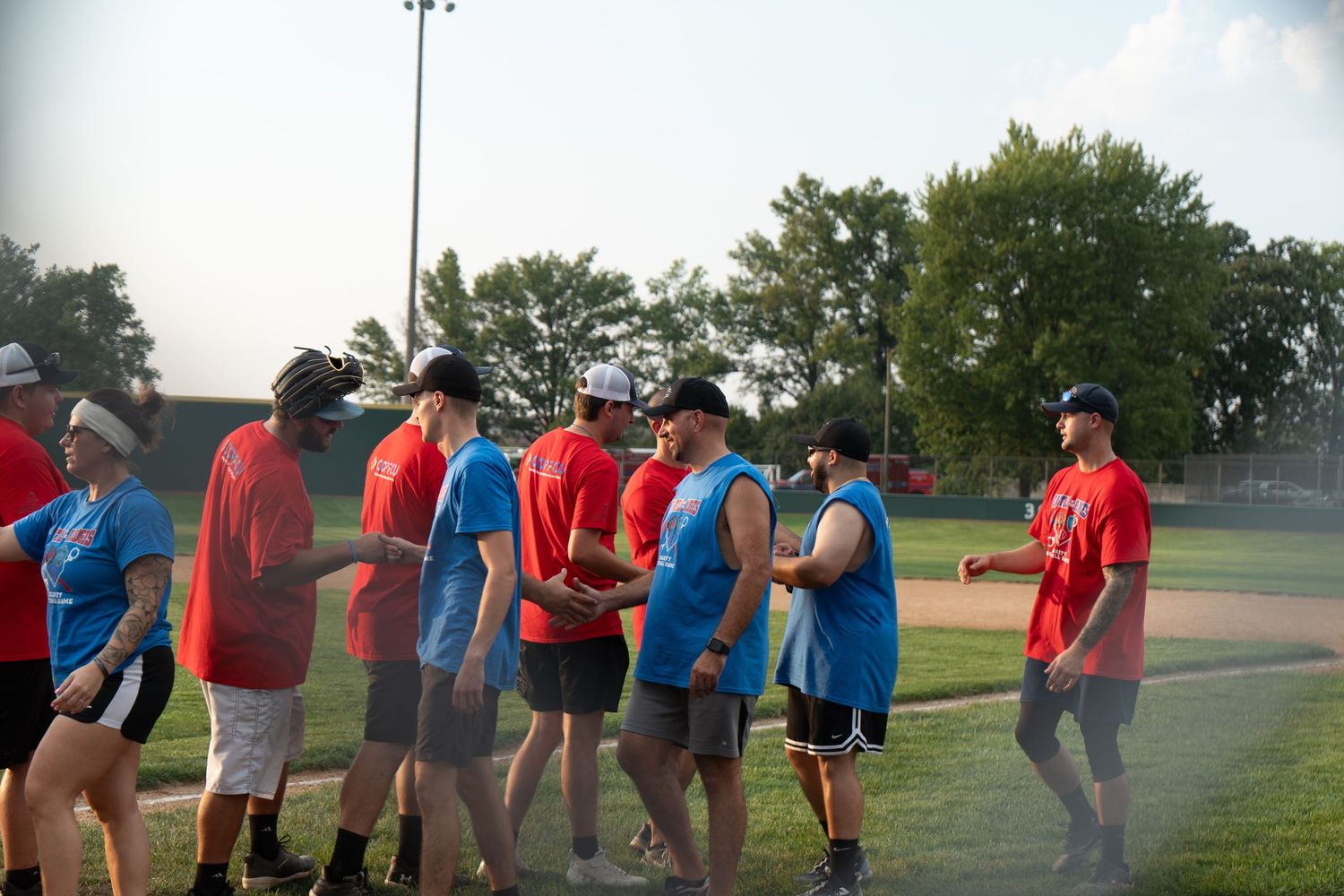 Teams shake hands on a baseball field after a recreational softball game on a sunny summer evening.