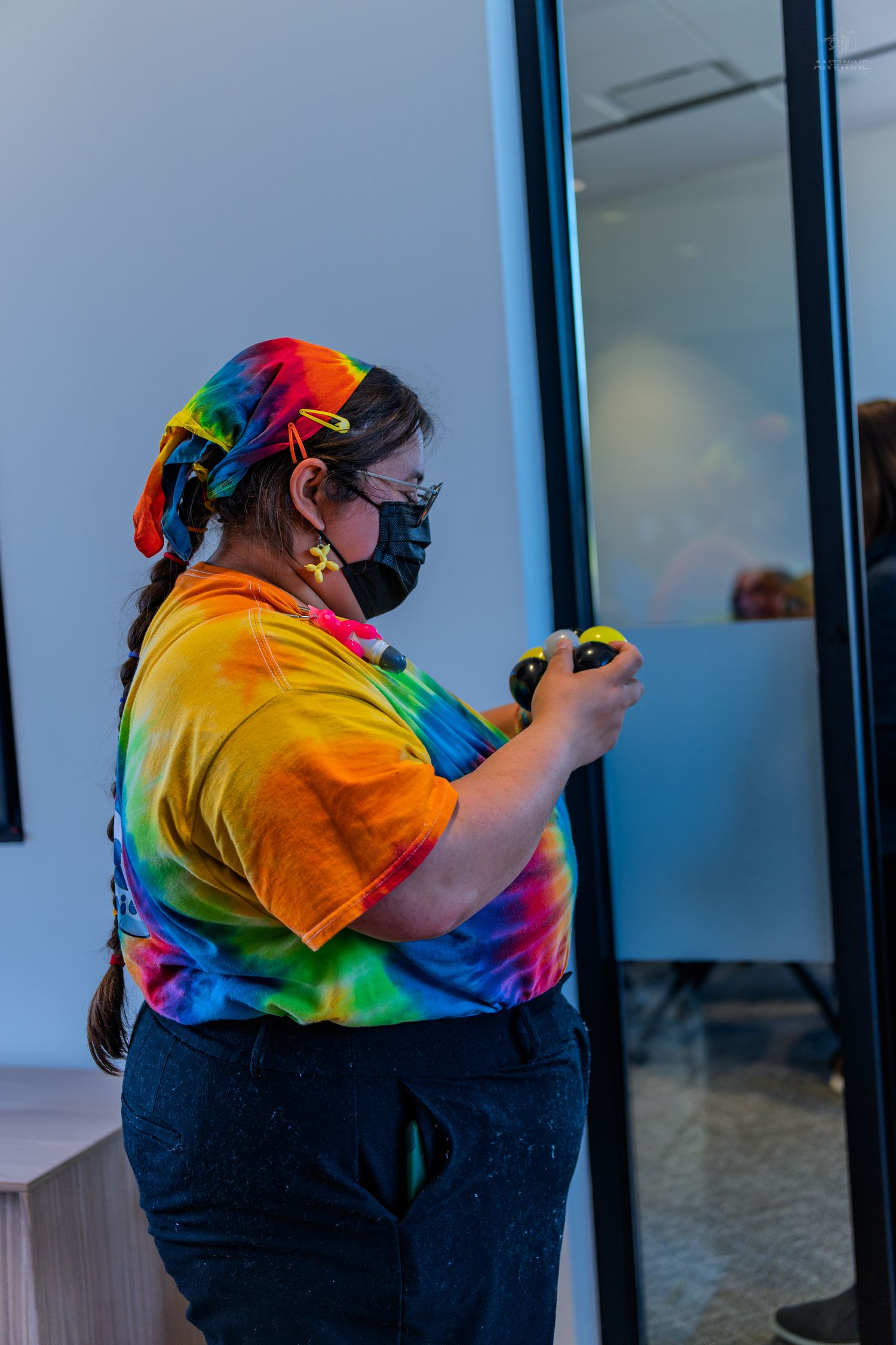 Person in colorful tie-dye shirt and black mask opening a glass door in an indoor setting.