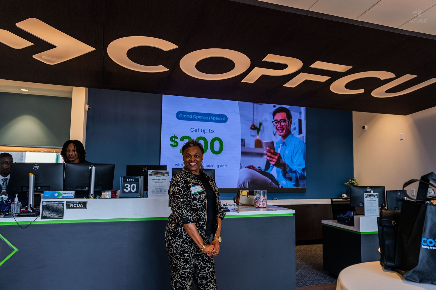 Woman smiling at COPFCU credit union branch counter with promotional display screen showing $200 offer.
