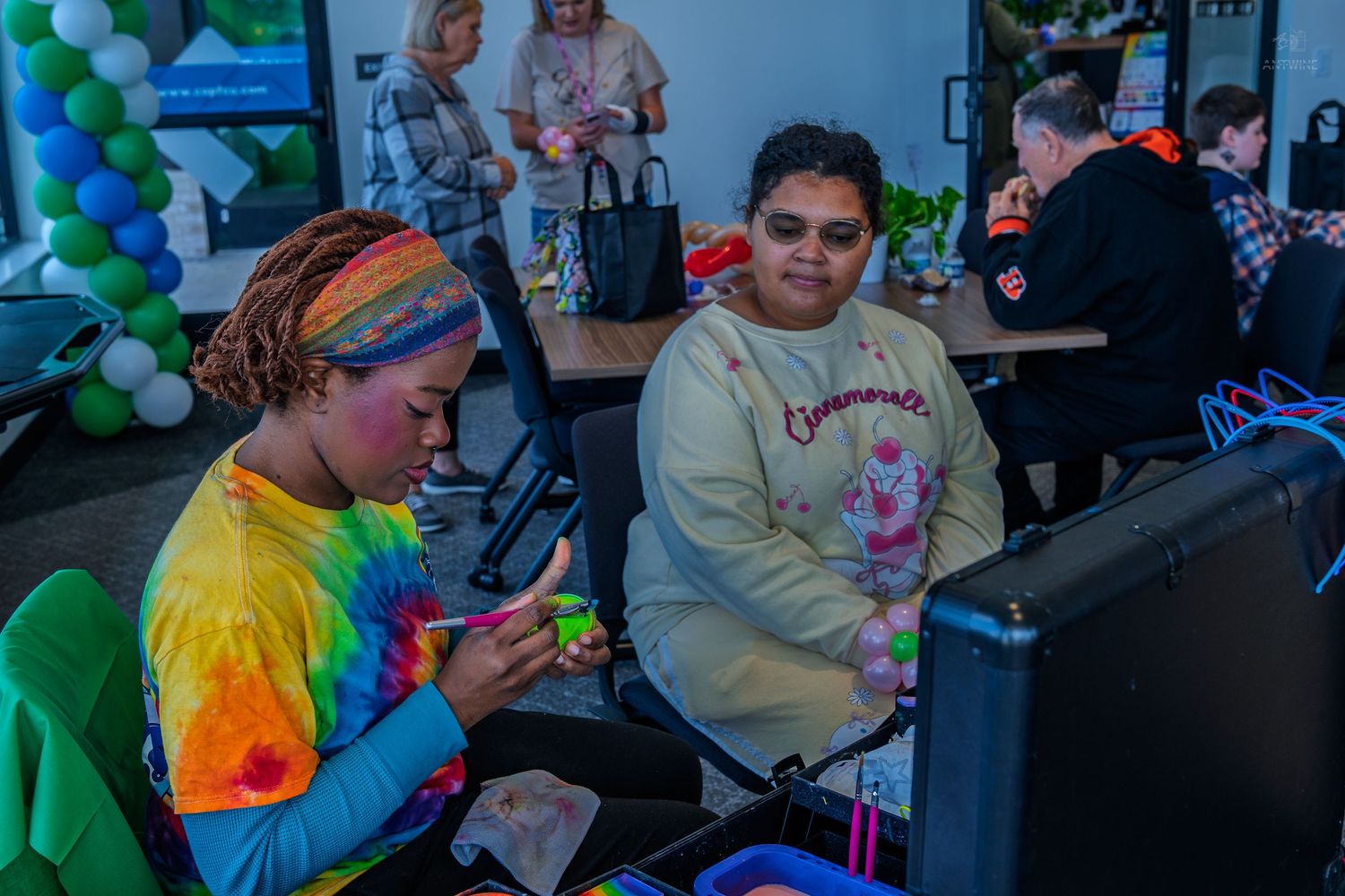 Two women in colorful attire work together at a table during a lively indoor craft event.